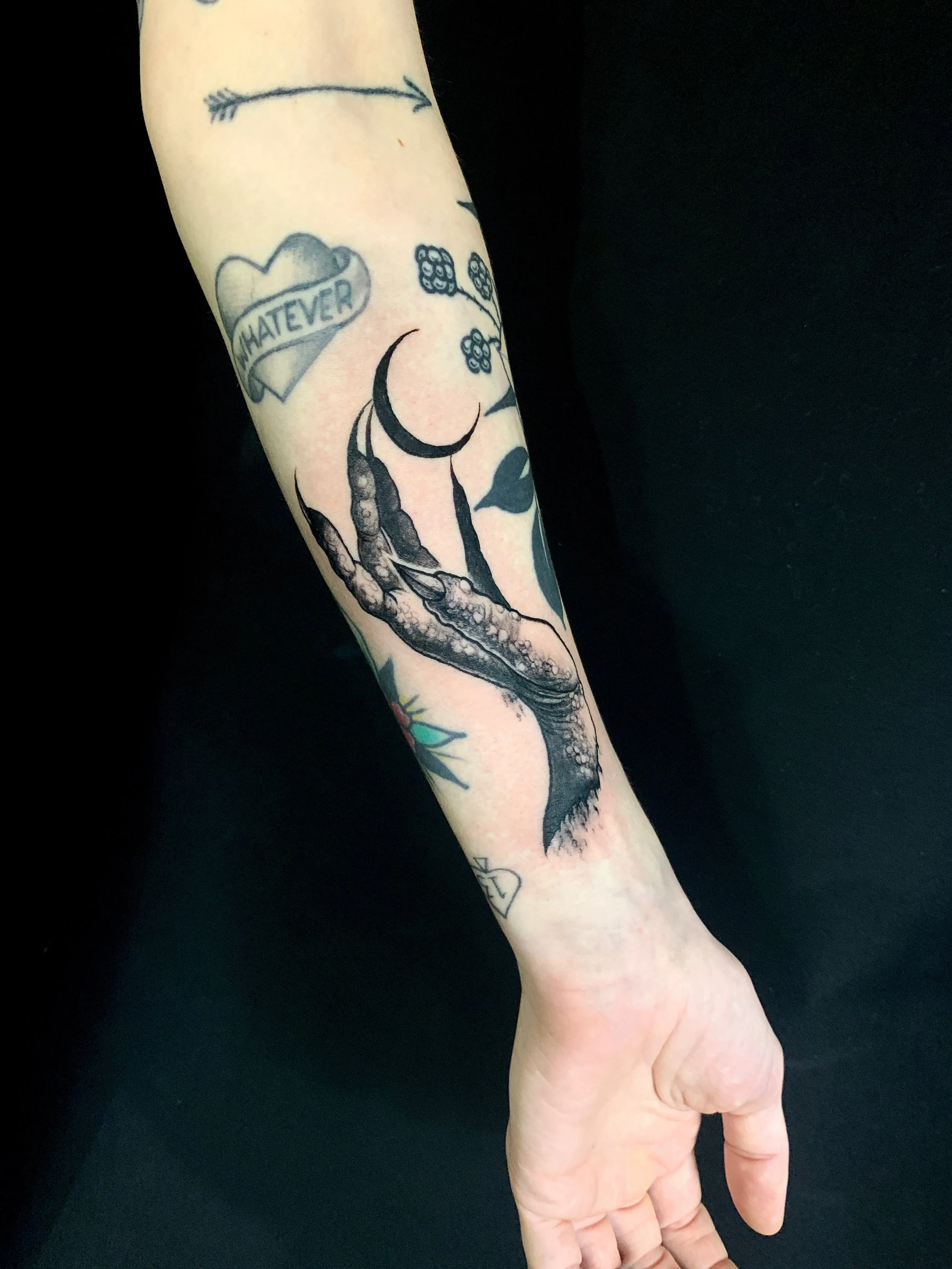 Tattoo of a skeletal hand holding a crescent moon on a person's forearm, surrounded by various smaller tattoos including a heart with the word 'WHATEVER', a cluster of small flowers or berries, and a small diamond shape.