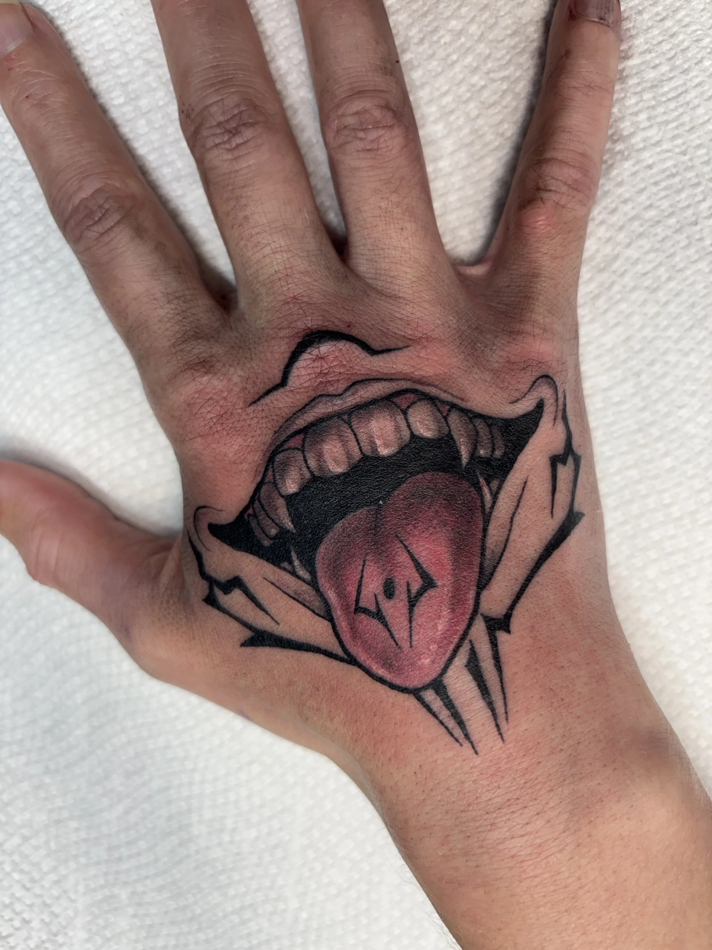 Tattoo of a mouth with sharp teeth and an open tongue, on the back of a hand.
