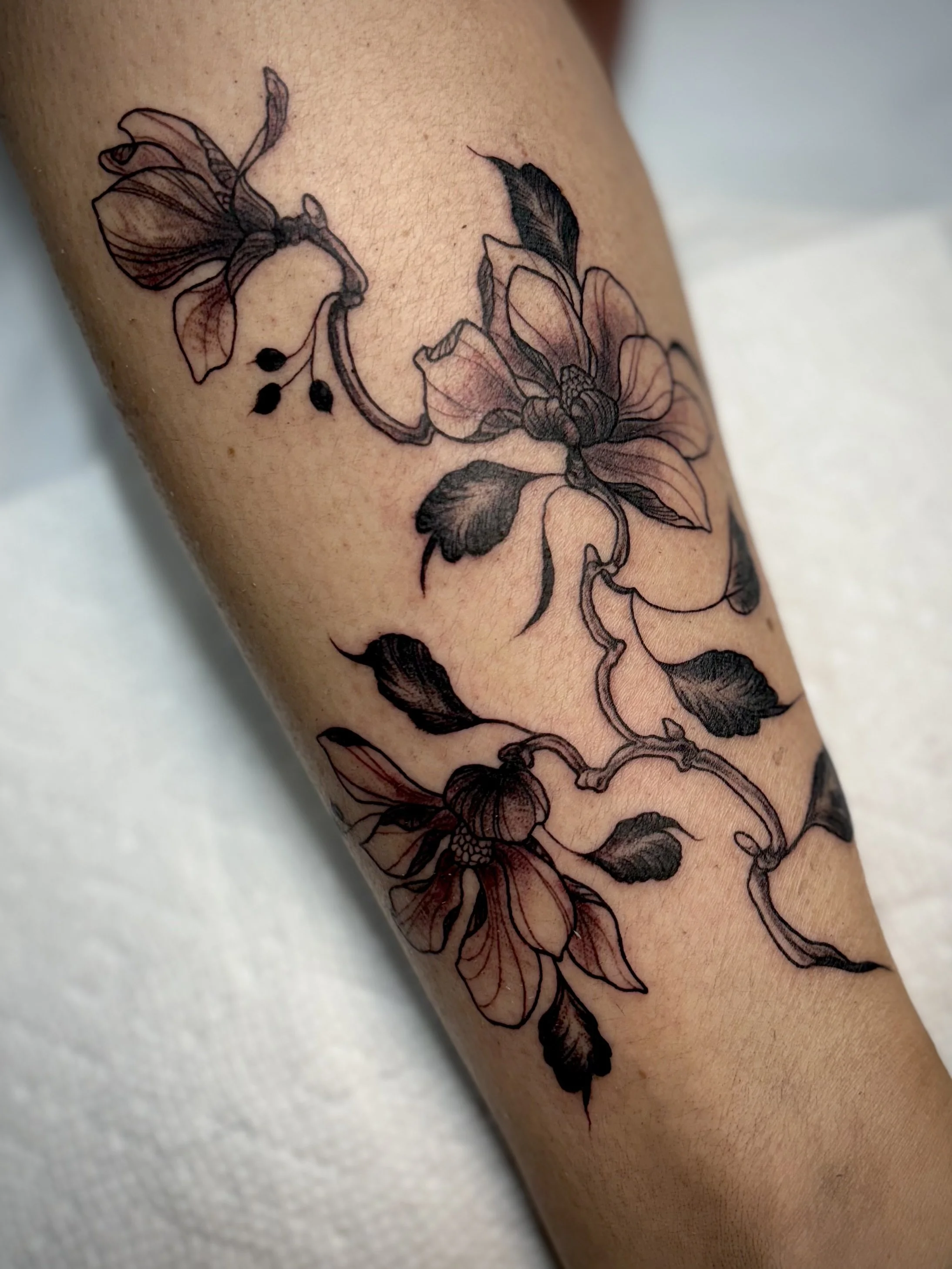 A black and gray floral tattoo featuring blooming magnolias, leaves, and twisting branches on a person's calf.