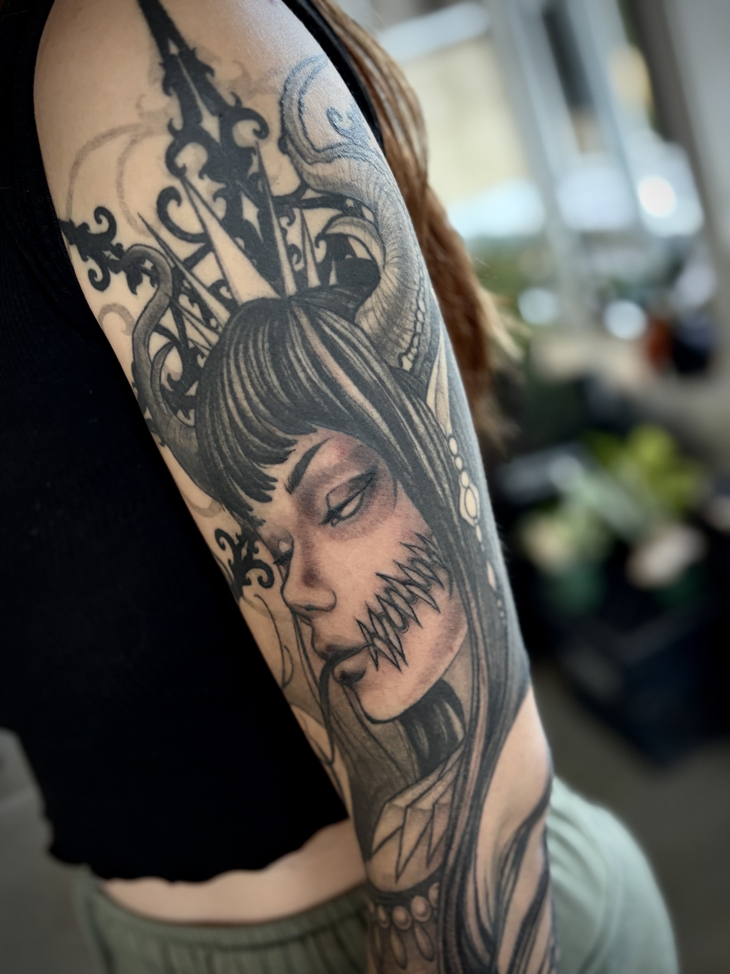 Tattoo of a demon woman with dark hair, horns, and vacant eyes, with a crown of spiked and an ornate background on a person's upper arm.