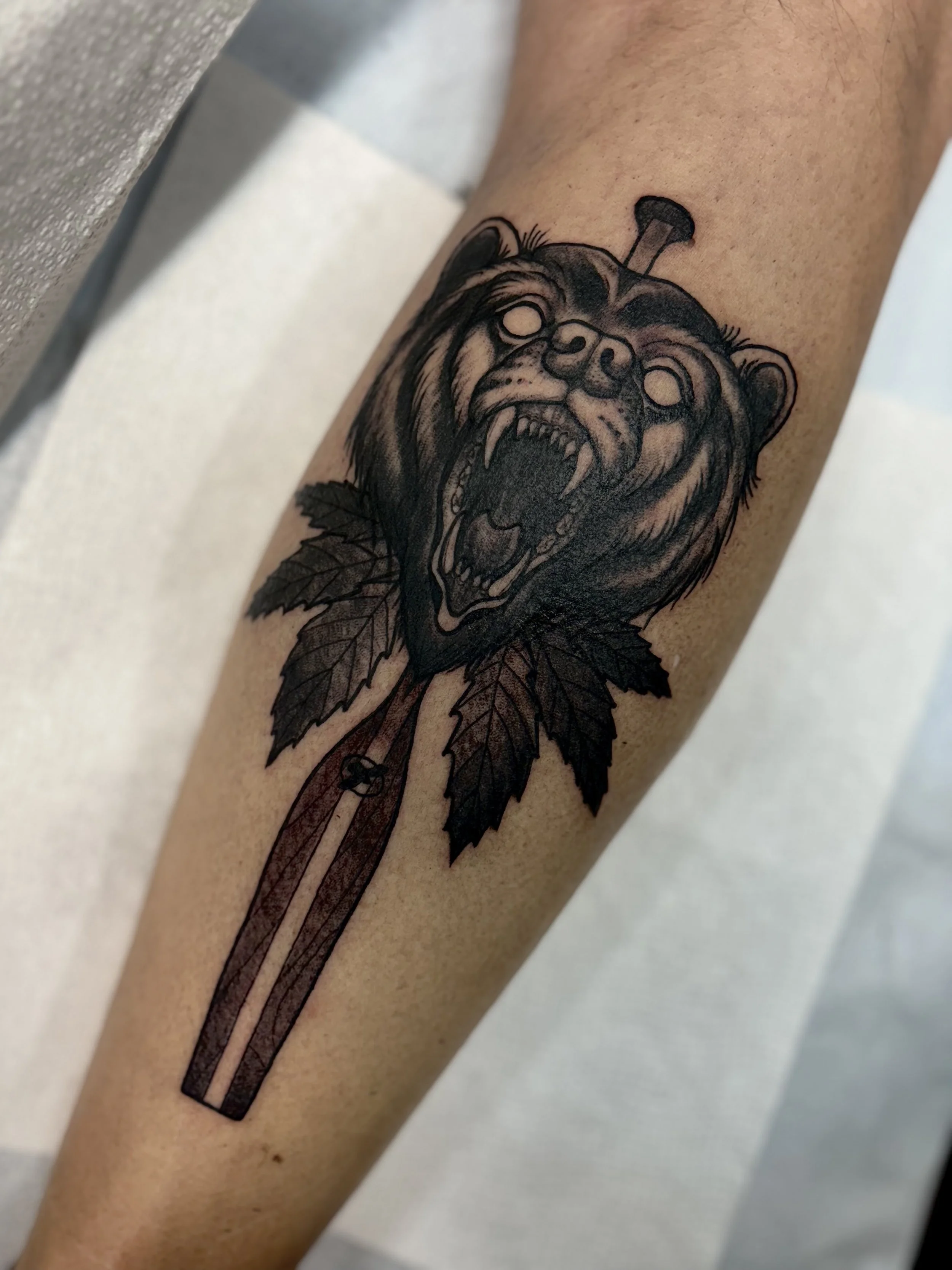 Tattoo of a bear's head with an open mouth and sharp teeth, surrounded by leaves, on a person's calf with a canoe paddle behind it.