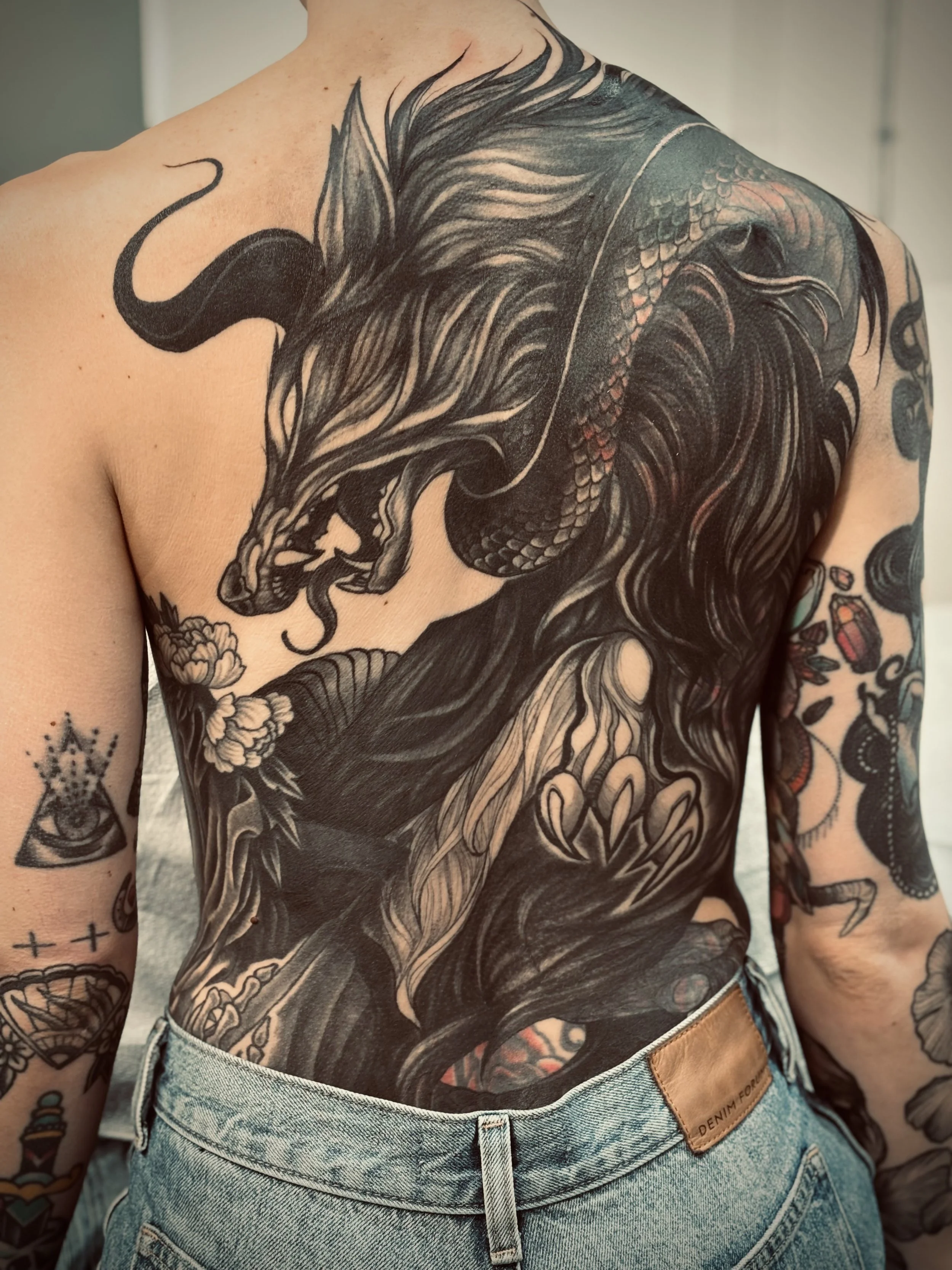 A person's back is covered with a large black and gray wolf and snake tattoo, featuring a fierce wolf with flowing fur fighting a large snake with detailed scales.