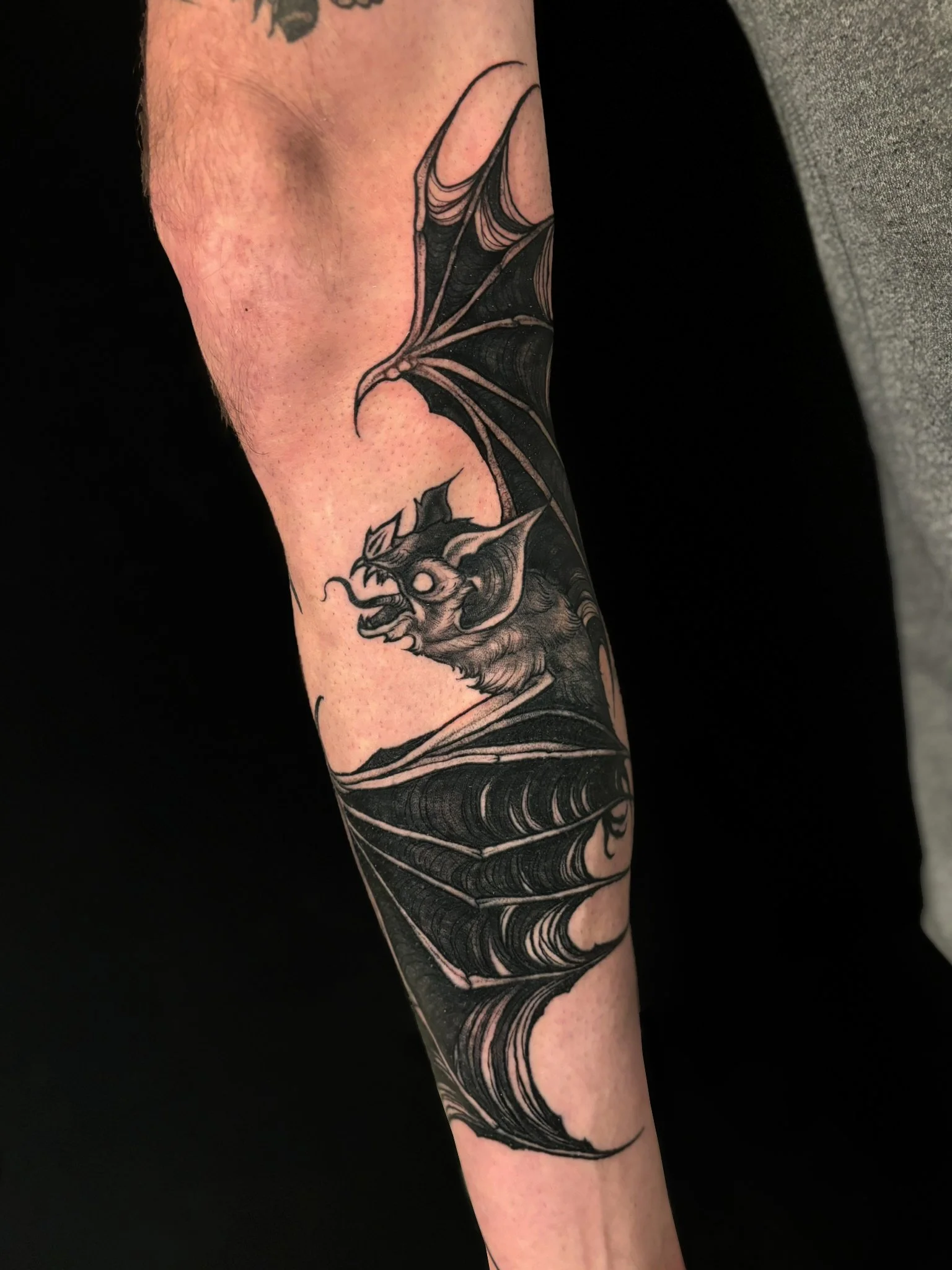 Tattoo of a bat with large wings on a person's leg, depicted in black ink with detailed shading.