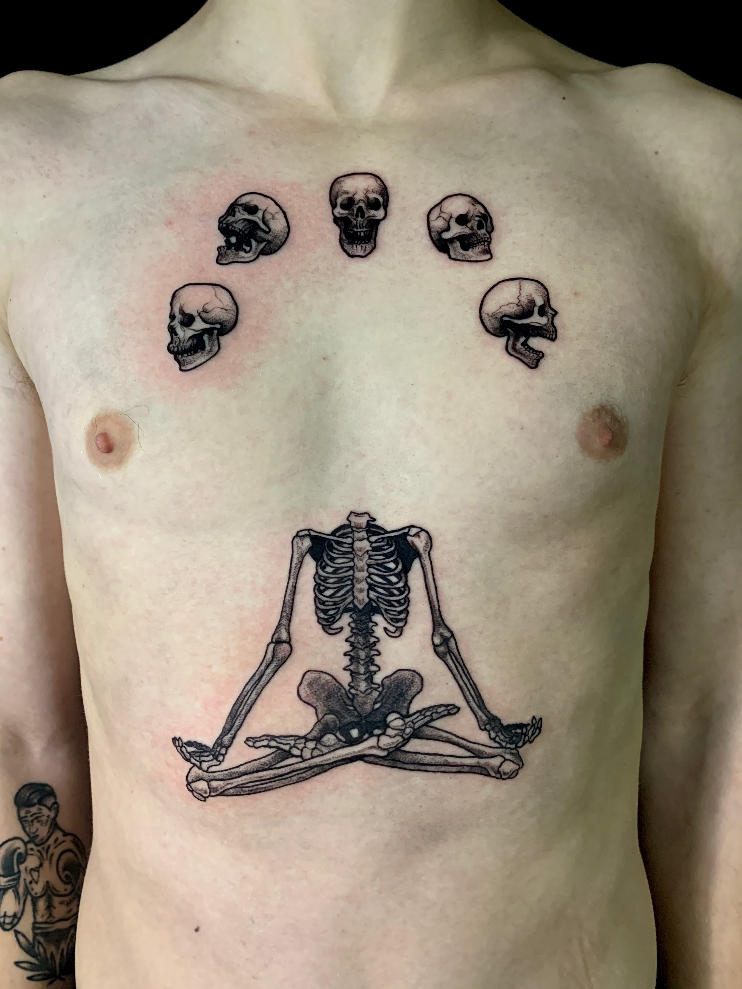 Black and grey tattoo of five skulls arranged in a semi-circle above a skeleton sitting in a cross-legged meditation pose.