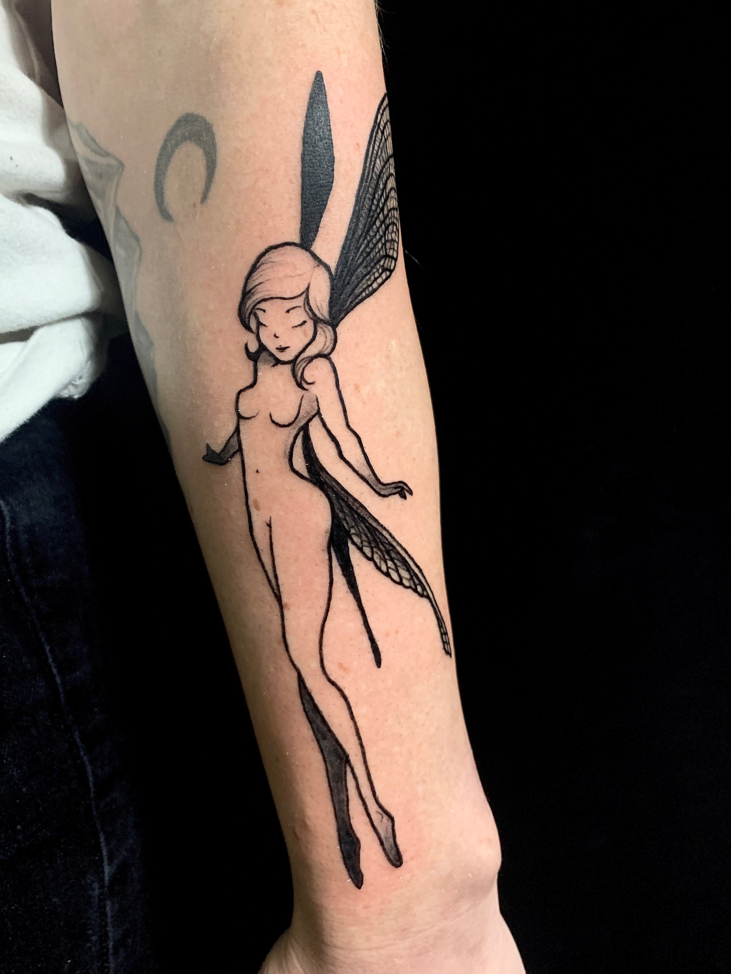Black and grey tattoo of a fairy resembling tinker bell with her hair down, and detailed dragonfly wings, on a person's forearm.