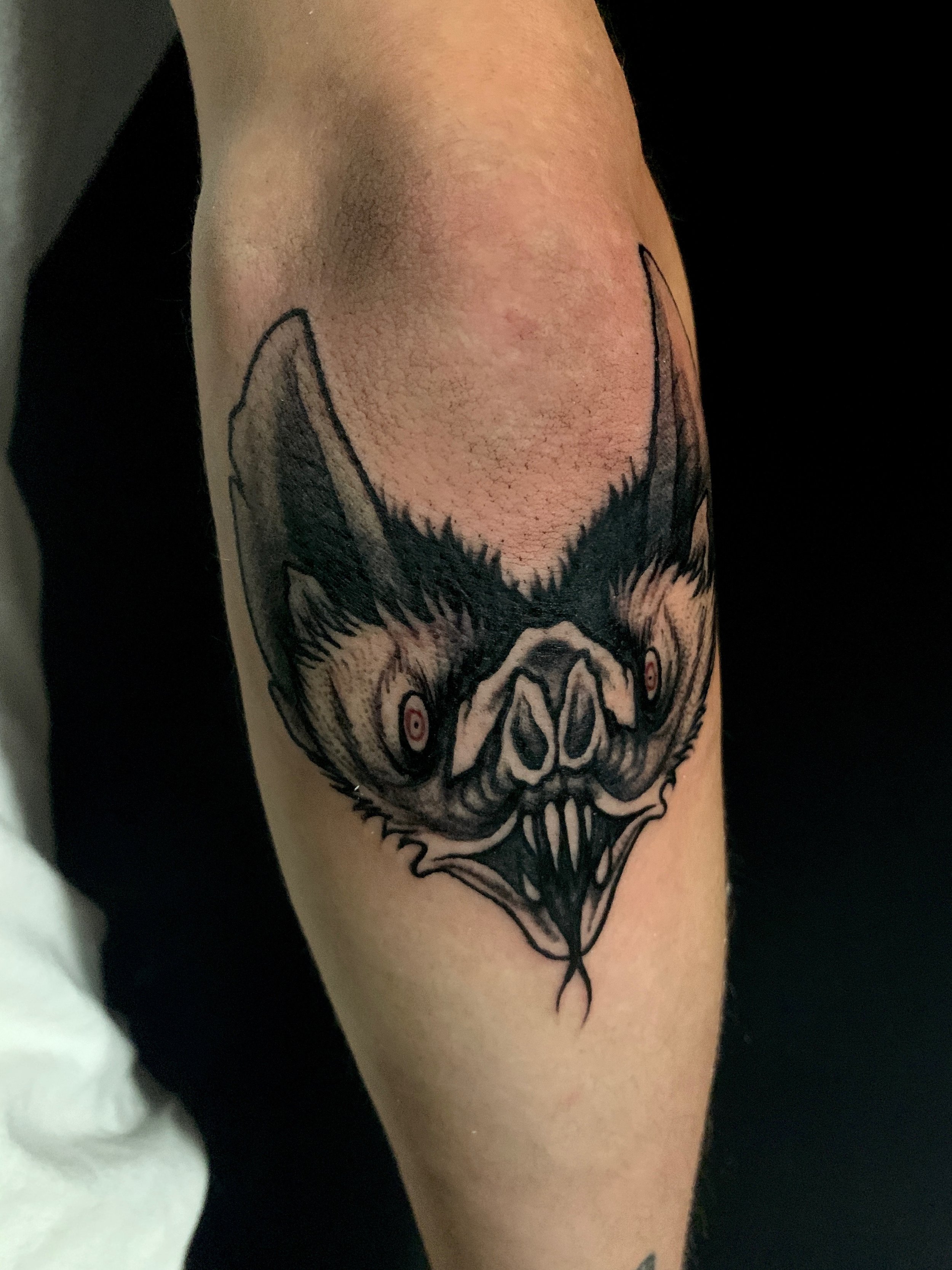 Tattoo of a menacing bat face with exaggerated facial features, sharp teeth, and red eyes on a person's lower arm near their elbow.