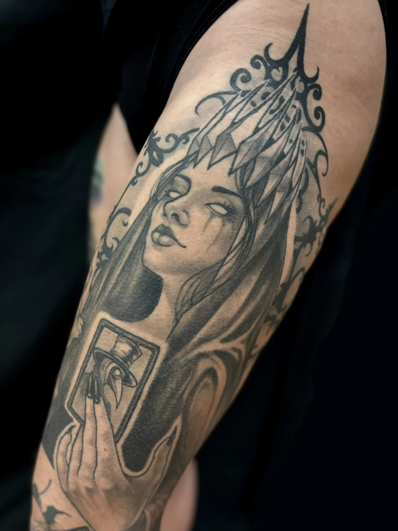 Black and gray tattoo on a person's thigh of a priestess woman's face with long flowing hair, holding a tarot card featuring a plague doctor, and wearing a crown of elongated spikes, surrounded by ornate decorative swirling patterns.