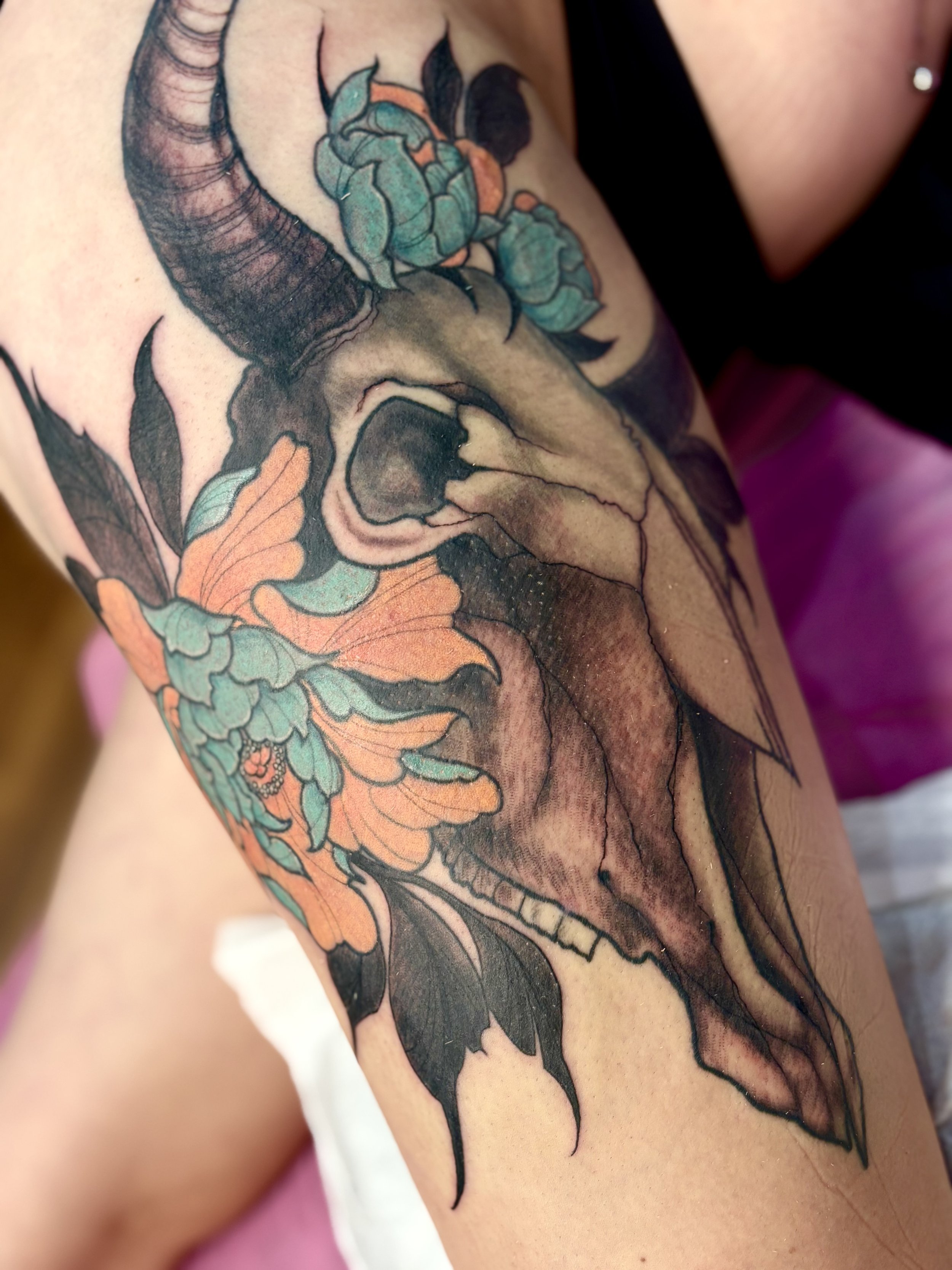 Tattoo of a long horn cow skull with floral and leaf design in black, gray, teal, and orange colors.