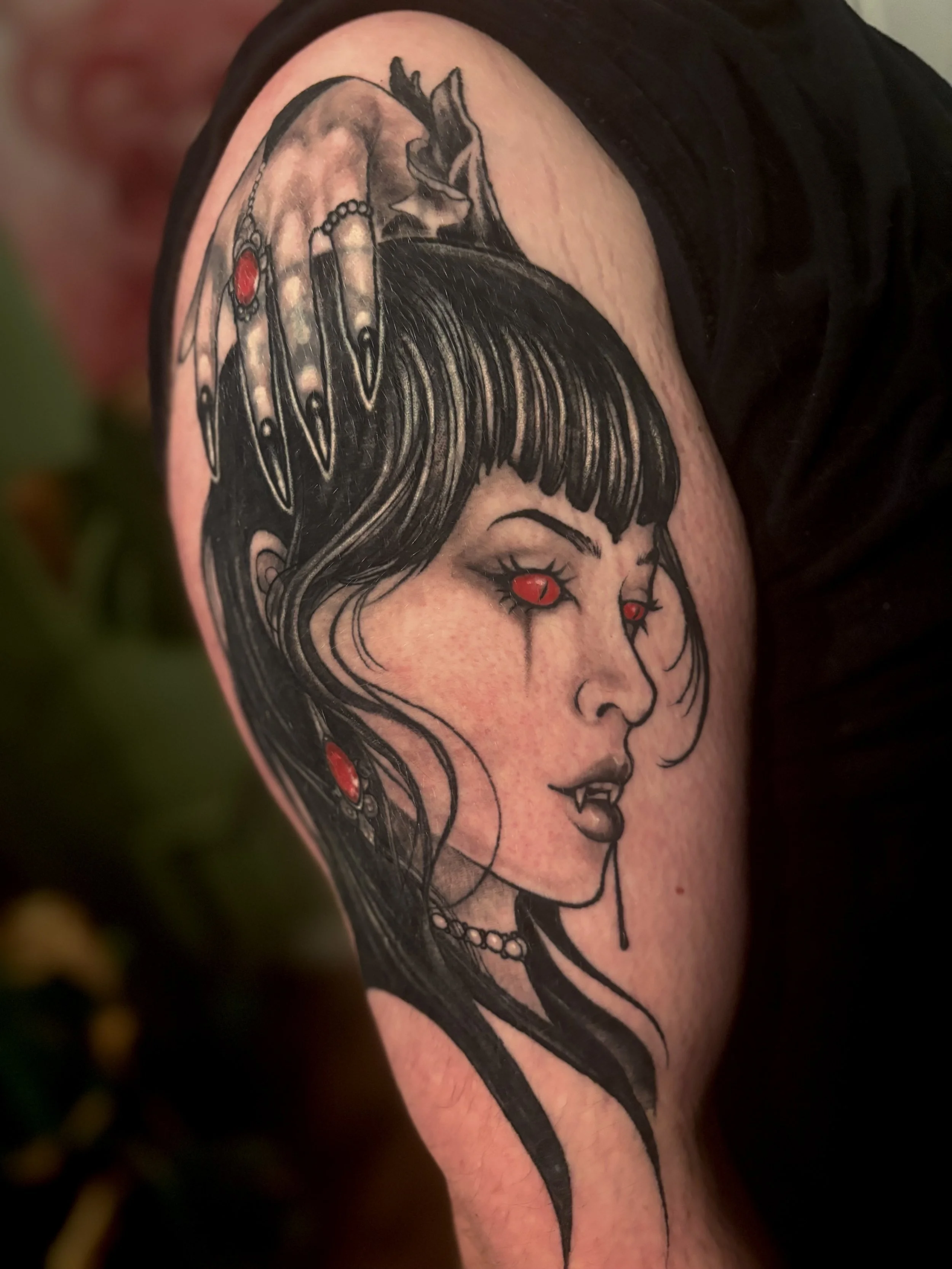 Tattoo of a vampire woman with black hair, red eyes, and fangs, depicted in a black and gray style with red highlights in her jewelry.