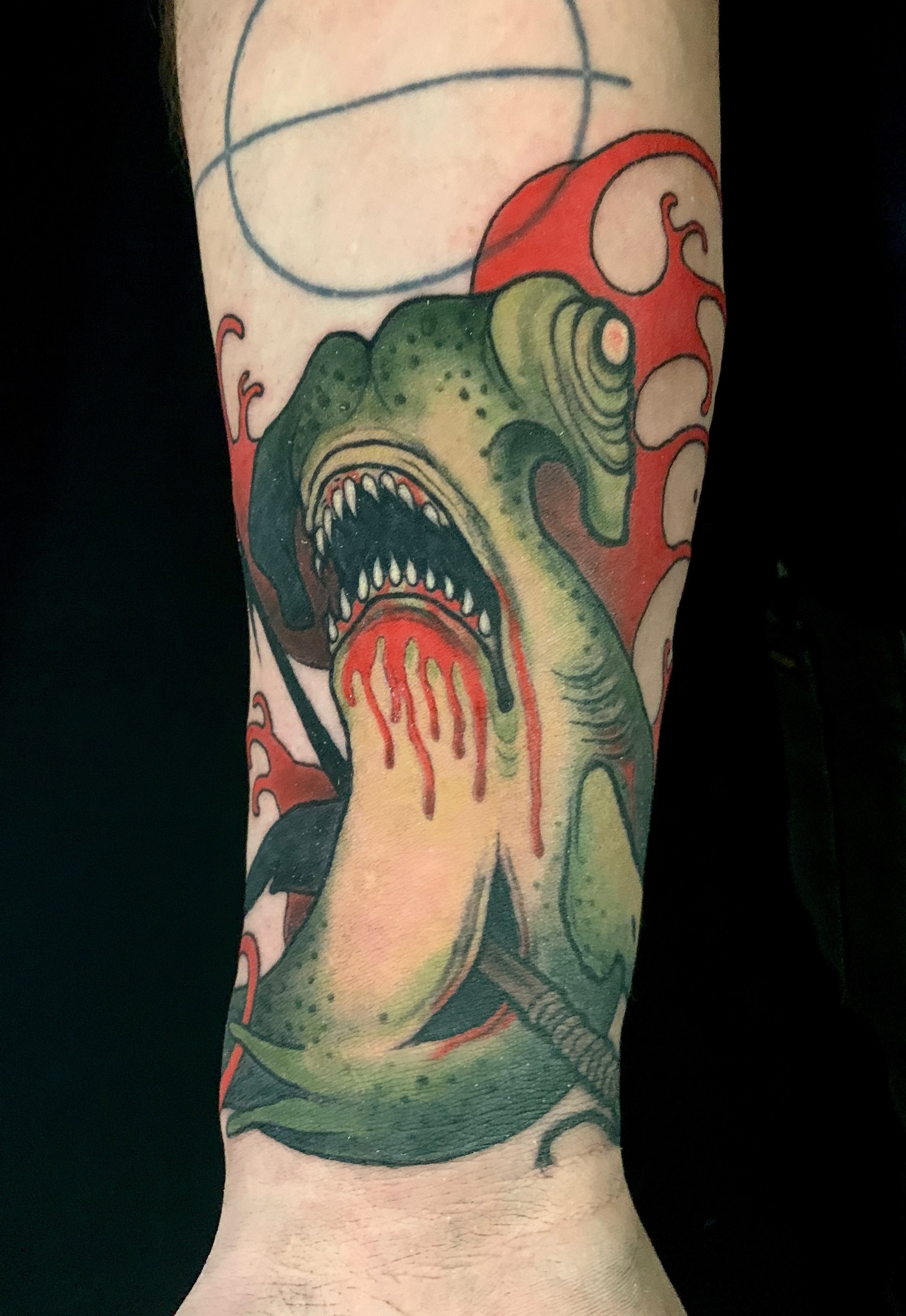 A tattoo of a zombie hammerhead shark with a large open mouth, sharp teeth, and greenish-yellow skin on a person's arm, with red and orange background elements.