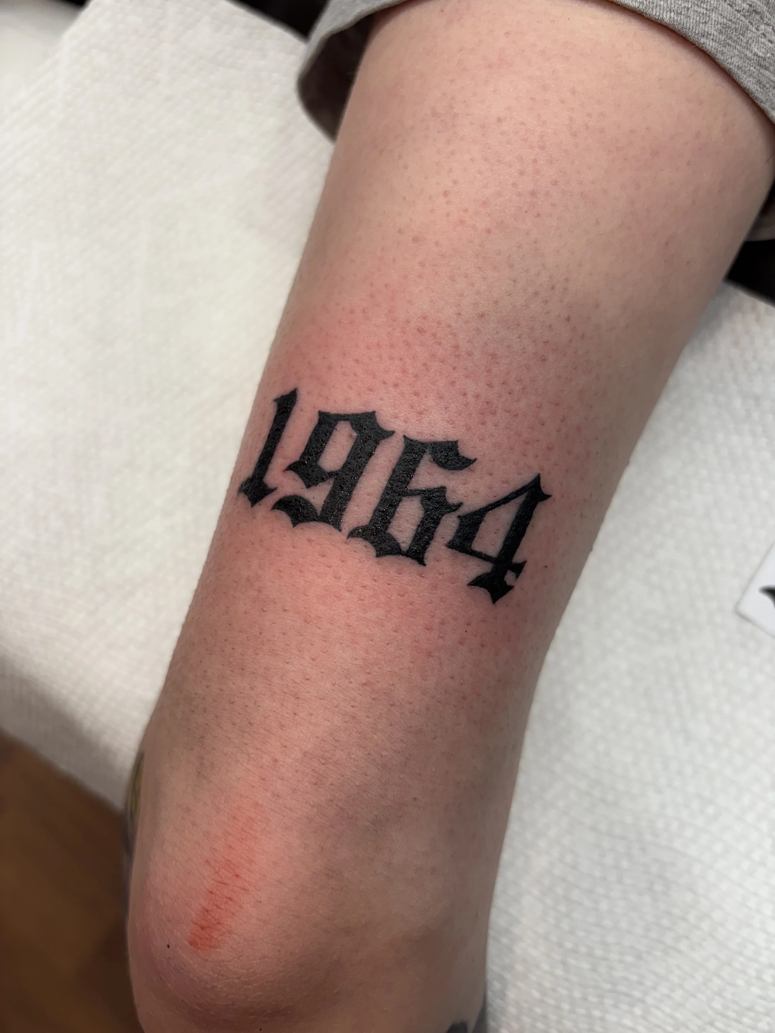 Tattoo of the year 1964 in black ink in a gothic font on a person's forearm.