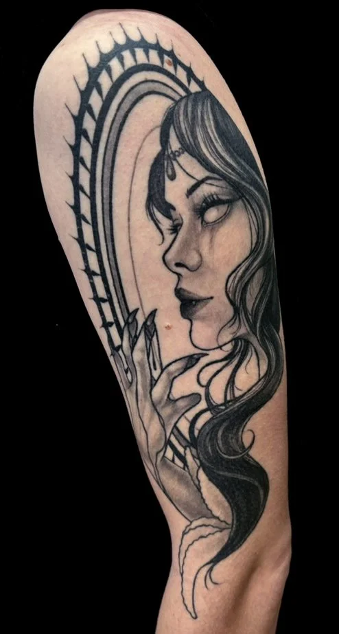 Black and gray tattoo of a vampire woman with long, wavy hair, and an ornamental crown on her forehead, holding her hand near her face, with an ornate, decorative background.