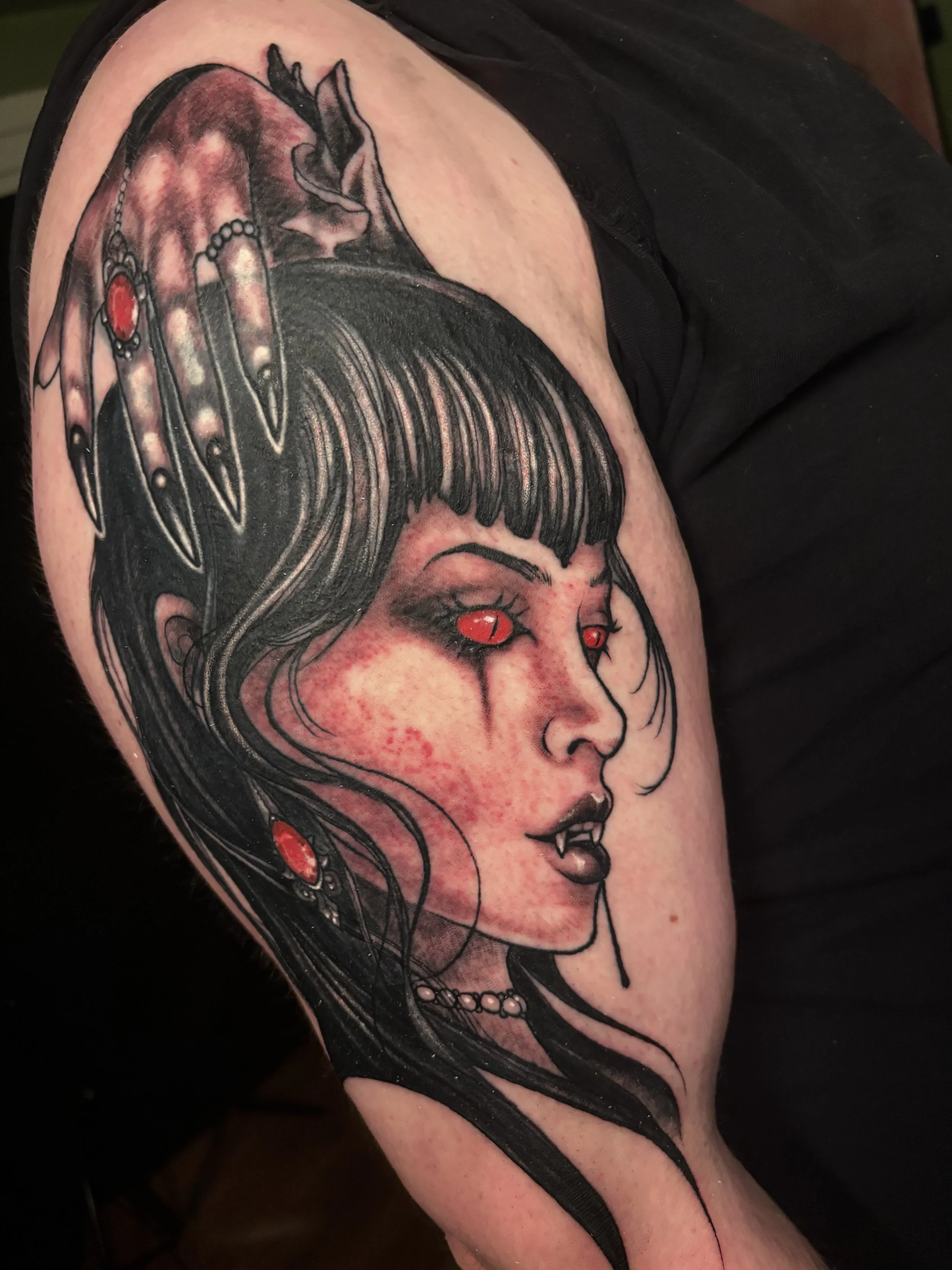 Tattoo of a woman with black hair, red eyes, vampire fangs, and tears of blood. She has black lipstick, and is wearing black jewelry with red gemstones.