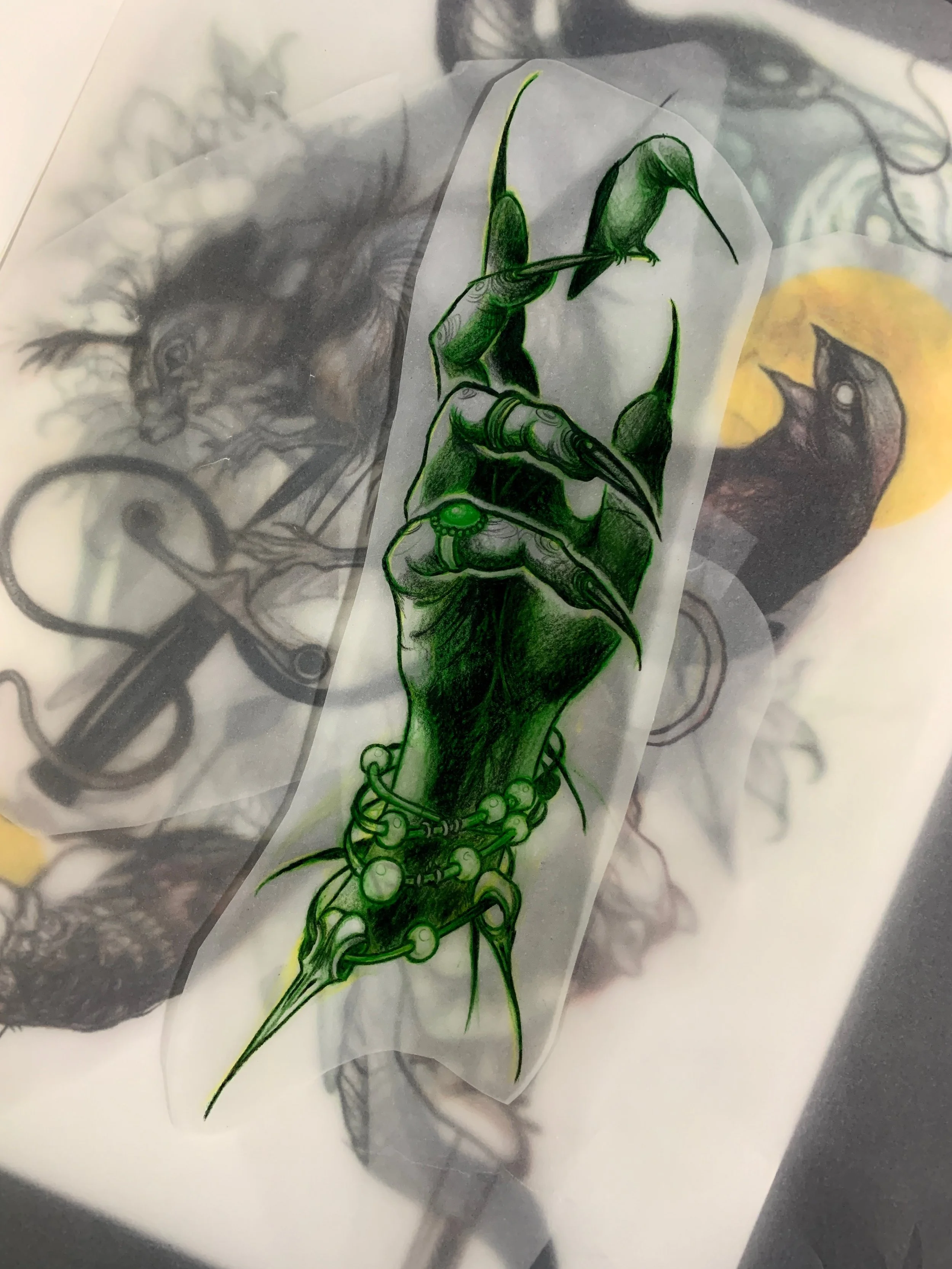 A green and black tattoo design of a hand with elongated, claw-like fingers, adorned with rings and a bracelet made of hummingbird skulls, and living hummingbird perched on its forefinger.