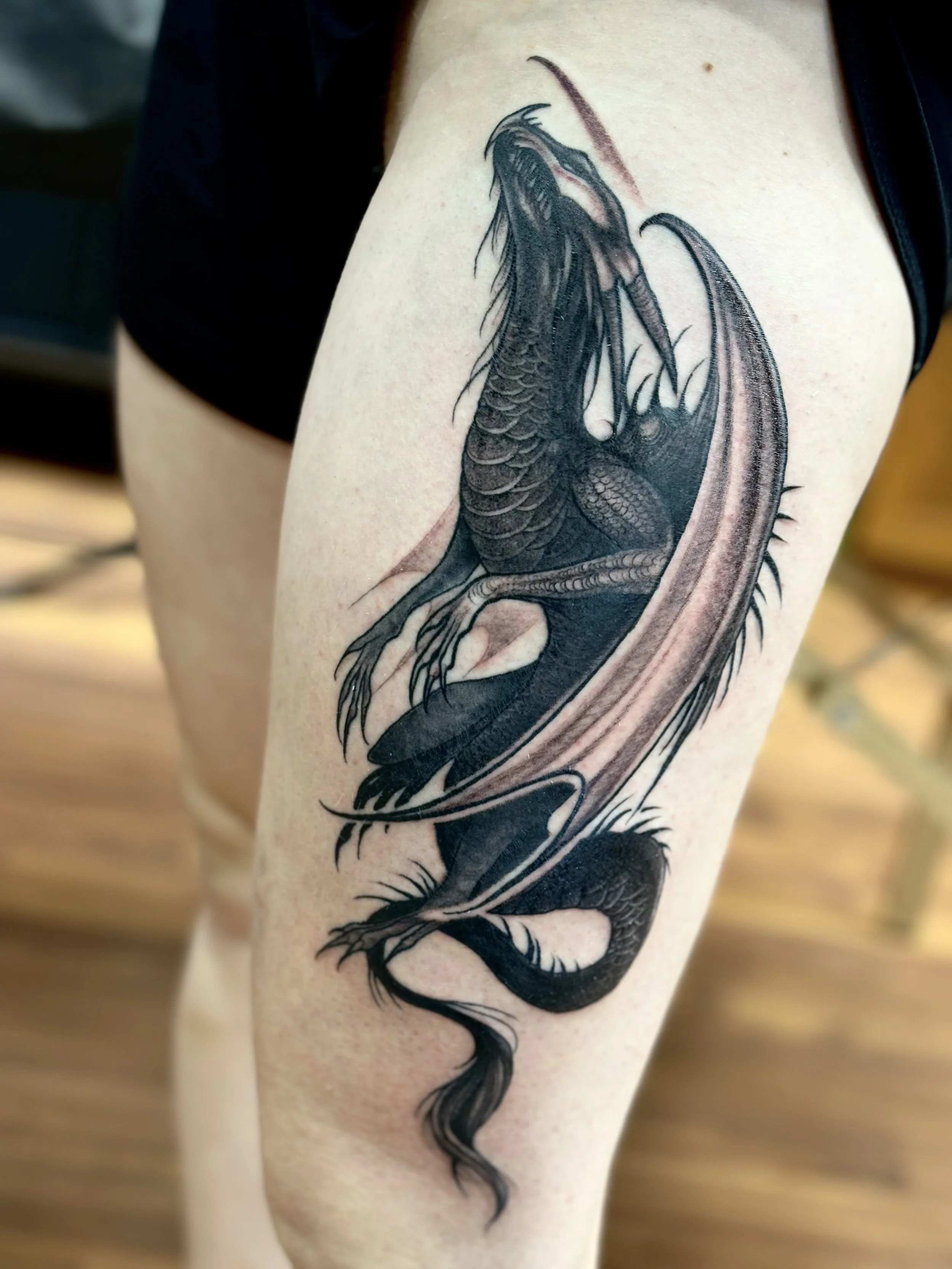 Black and grey tattoo of a dragon on a person's thigh.