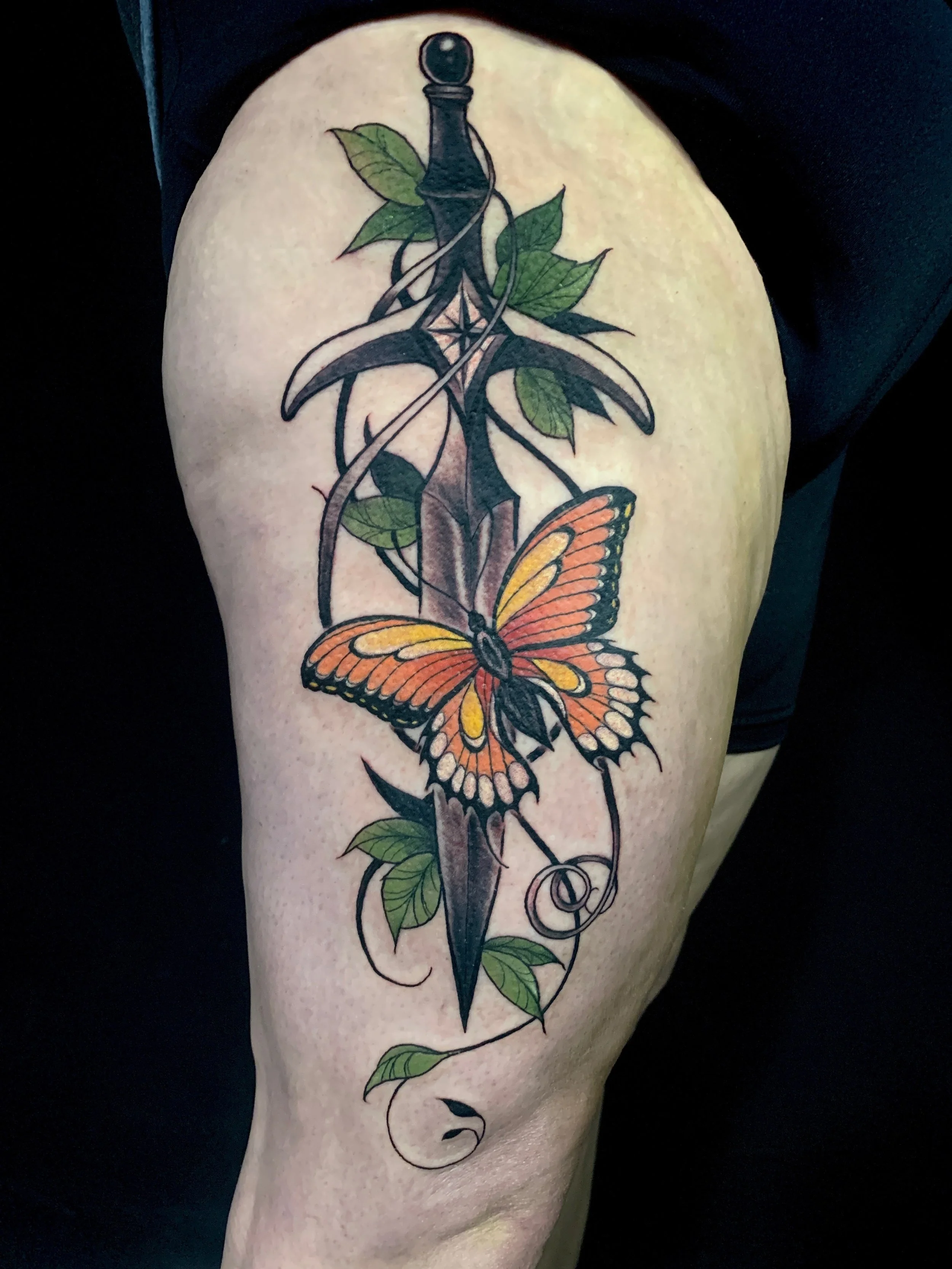 Tattoo of a butterfly with orange, yellow, and black wings on a person's thigh, intertwined with a dagger and green leaves.