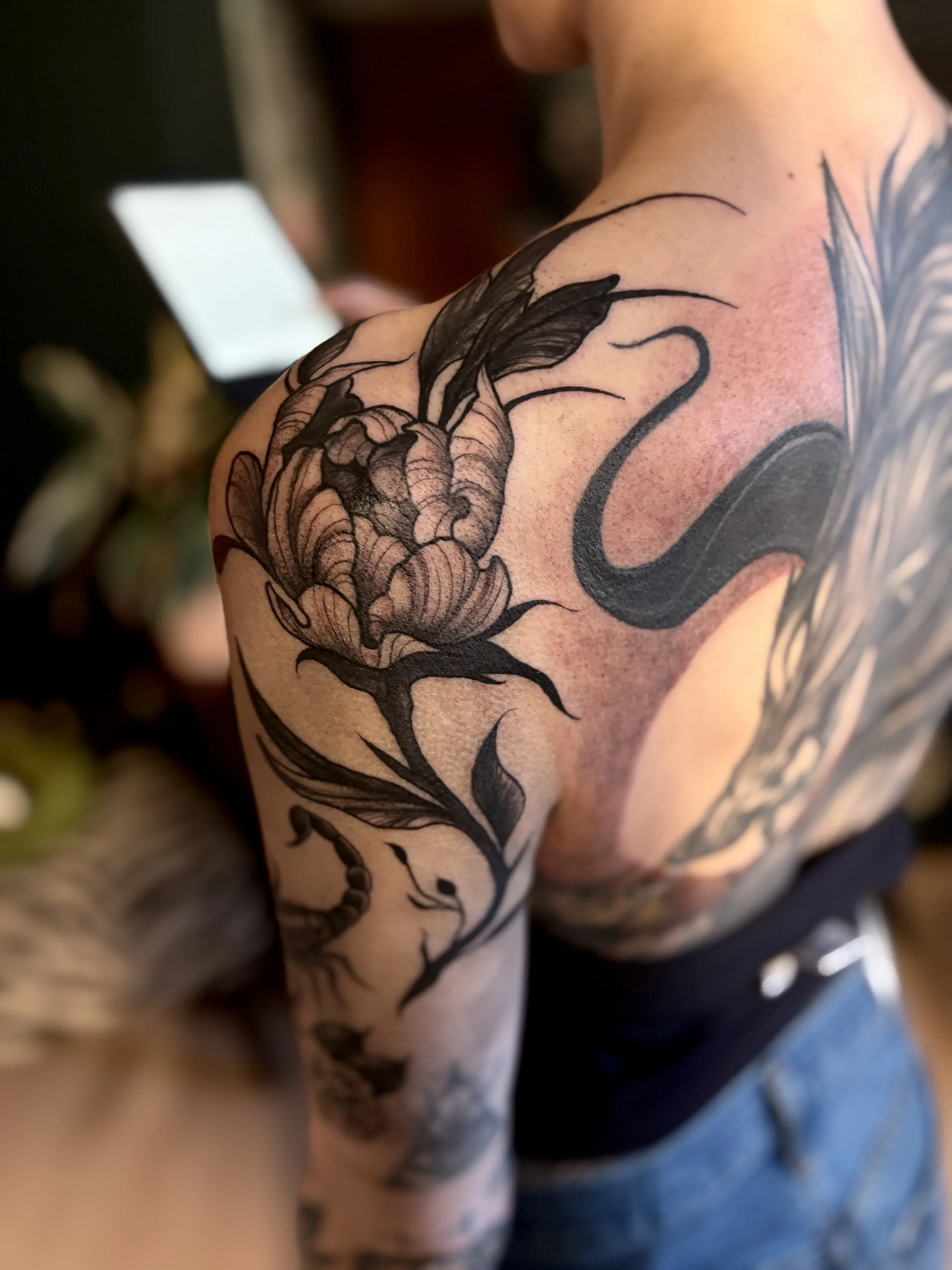 Close-up of a person's shoulder and upper arm with a detailed black tattoo of a peony, with swirling long leaves and stems.