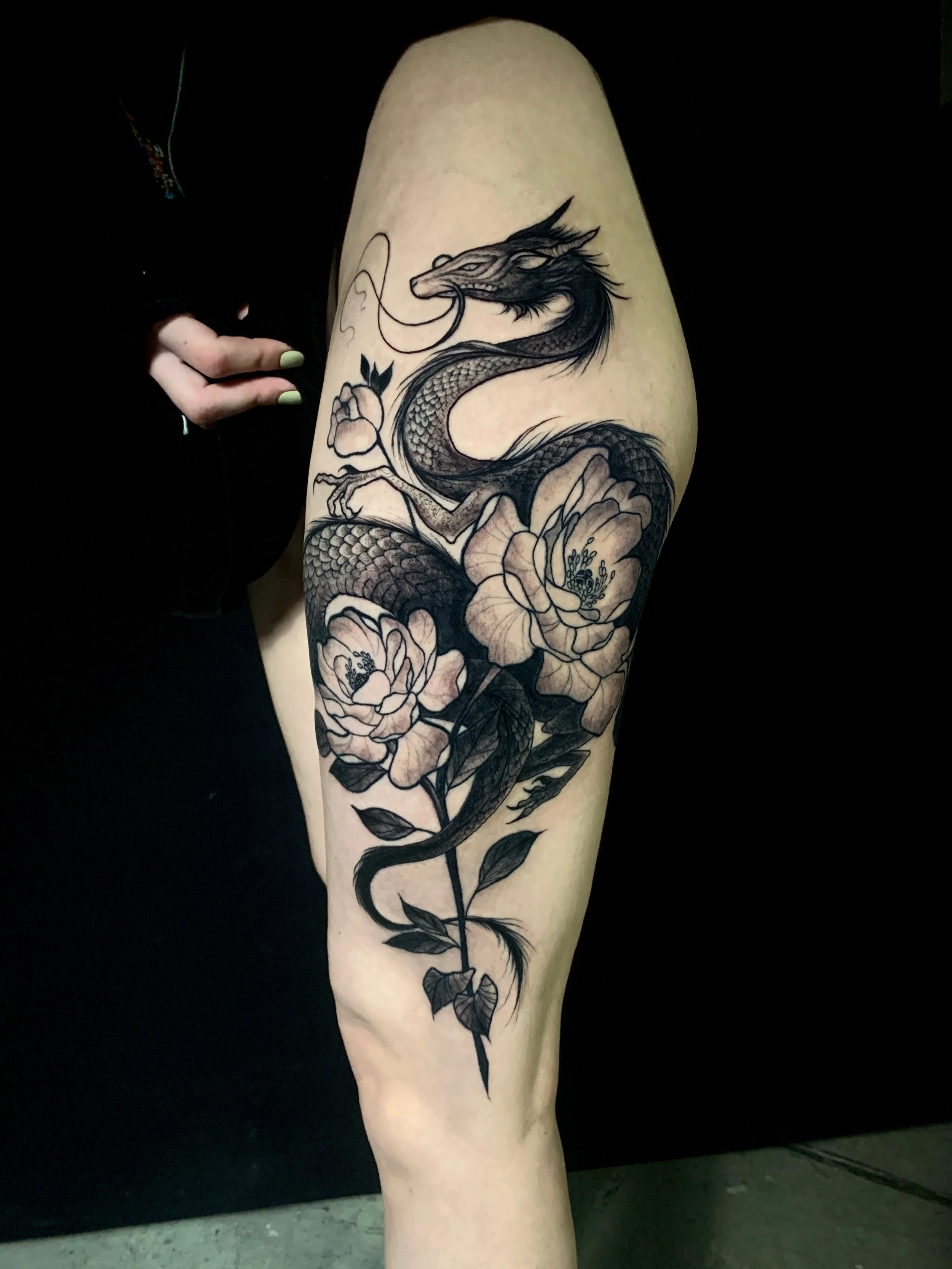 Black and gray tattoo of a dragon coiled around flowers on a person's thigh.