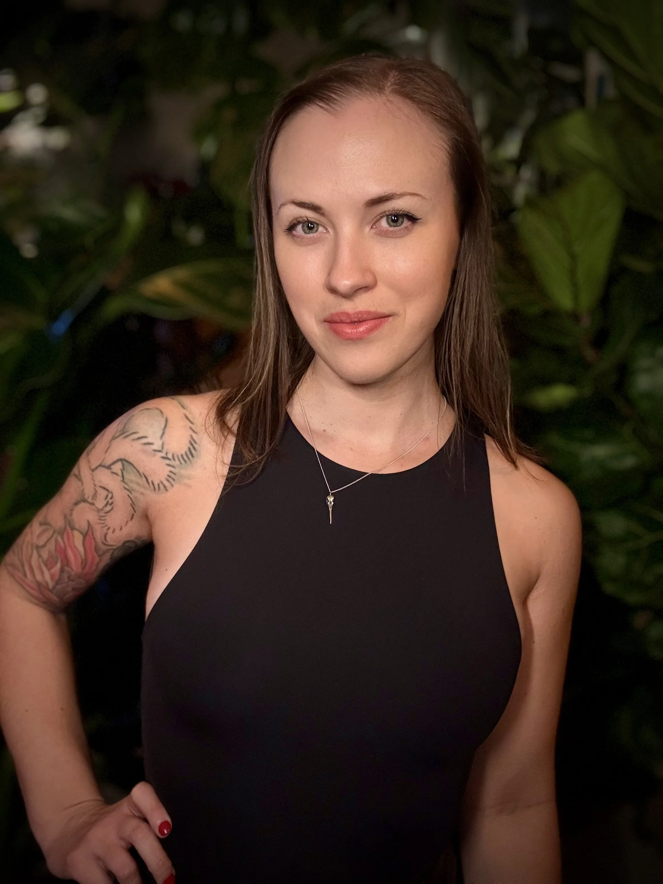 A woman with light skin, long brown hair, and blue eyes. She is wearing a sleeveless black top, a delicate necklace, and has a tattoo on her left shoulder. She is standing outdoors with a background of large green leaves.
