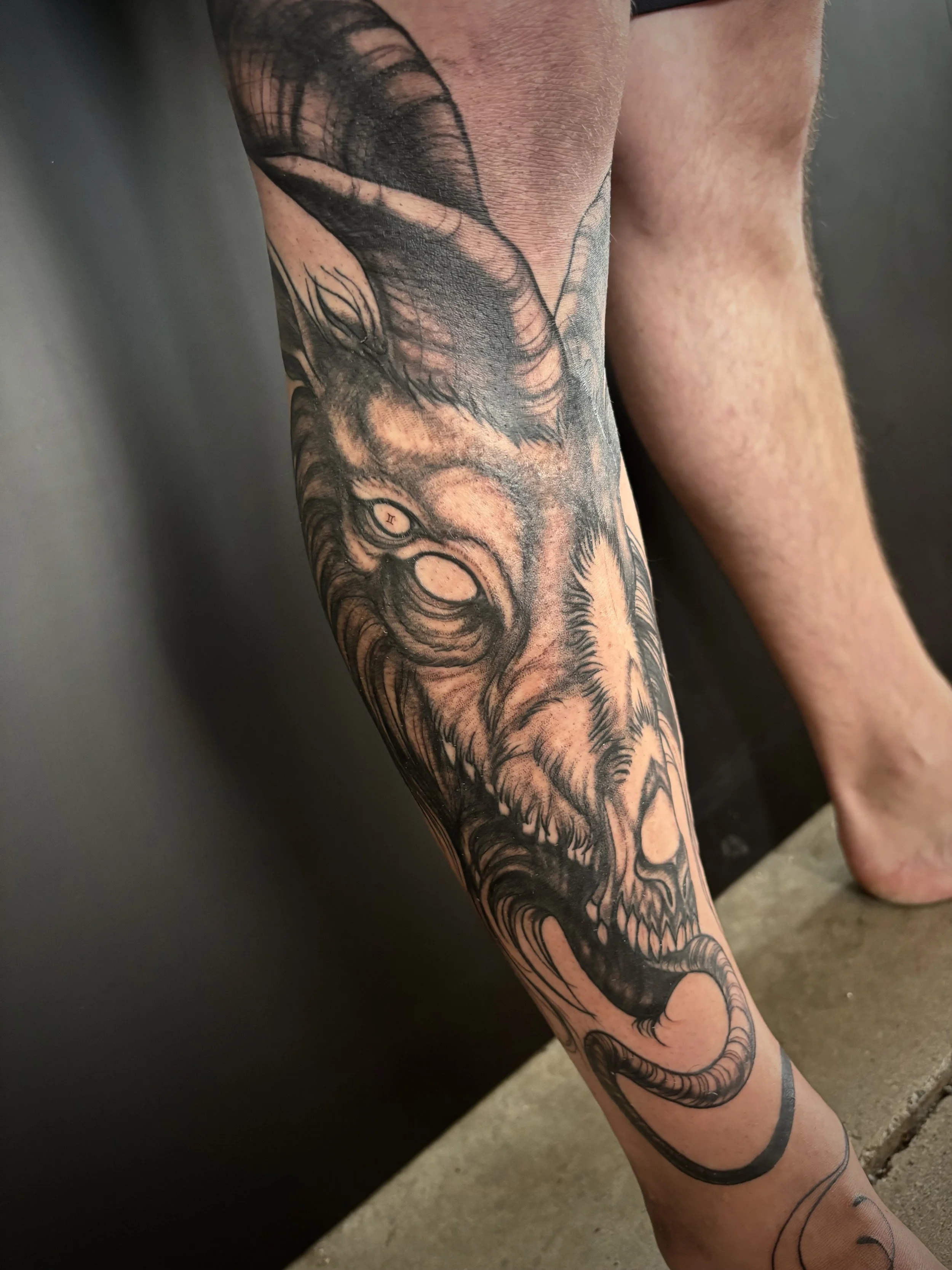 Black and gray tattoo of a demonic goat creature with horns, sharp teeth, a long tongue, and glowing vacant eyes on a person's leg.