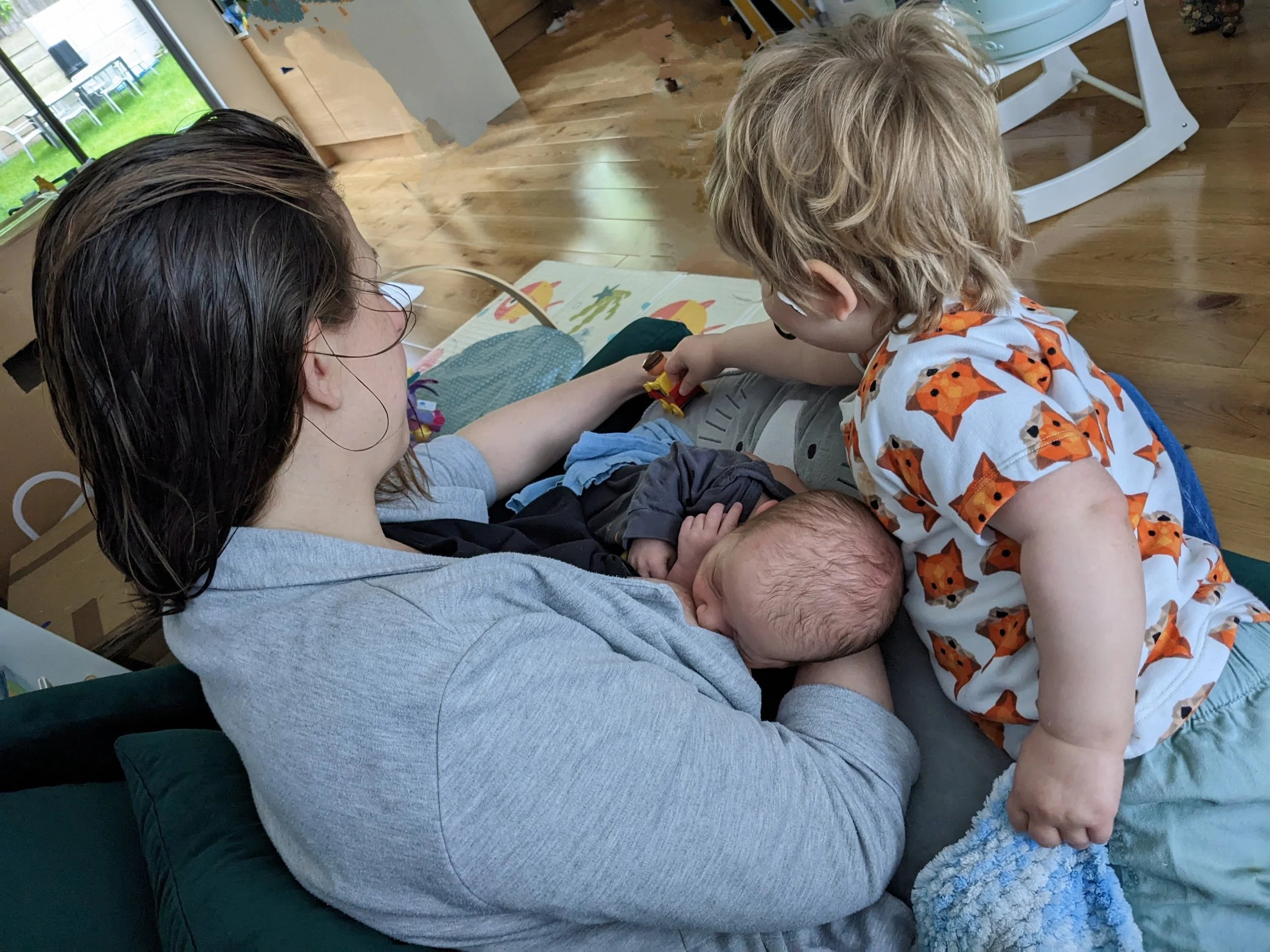 A happy mum feeding her new baby while her toddler plays beside her, depicting the joys of having a second child and balancing family life after Antenatal Classes in Walthamstow.