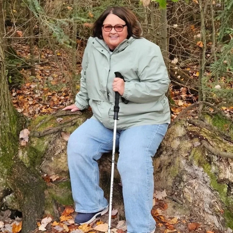 A woman sitting on a fallen tree trunk in a forest, wearing glasses, a light gray jacket, blue jeans, and holding a walking stick, surrounded by trees and fallen autumn leaves.