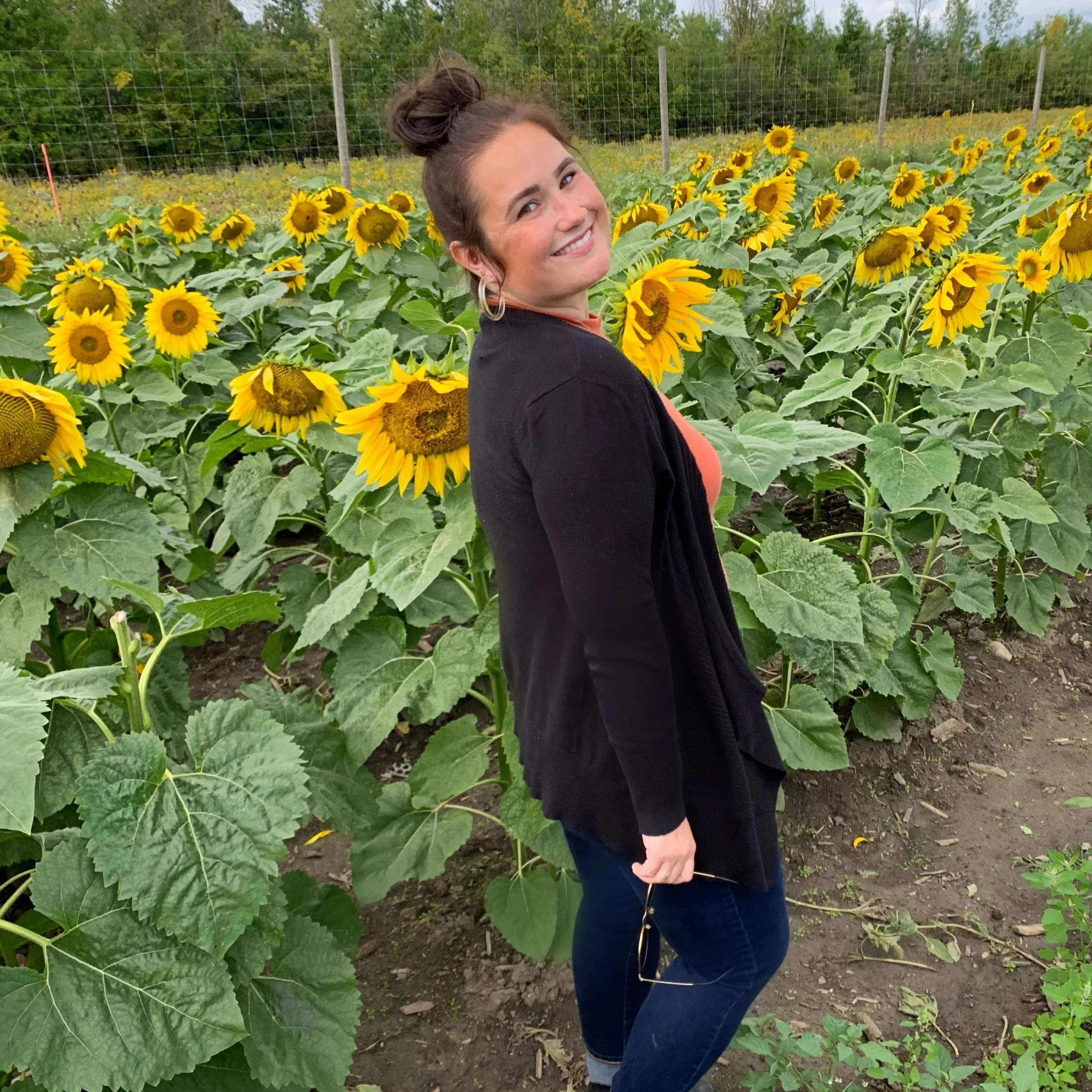 A woman smiling and standing among blooming sunflowers in a sunflower field.