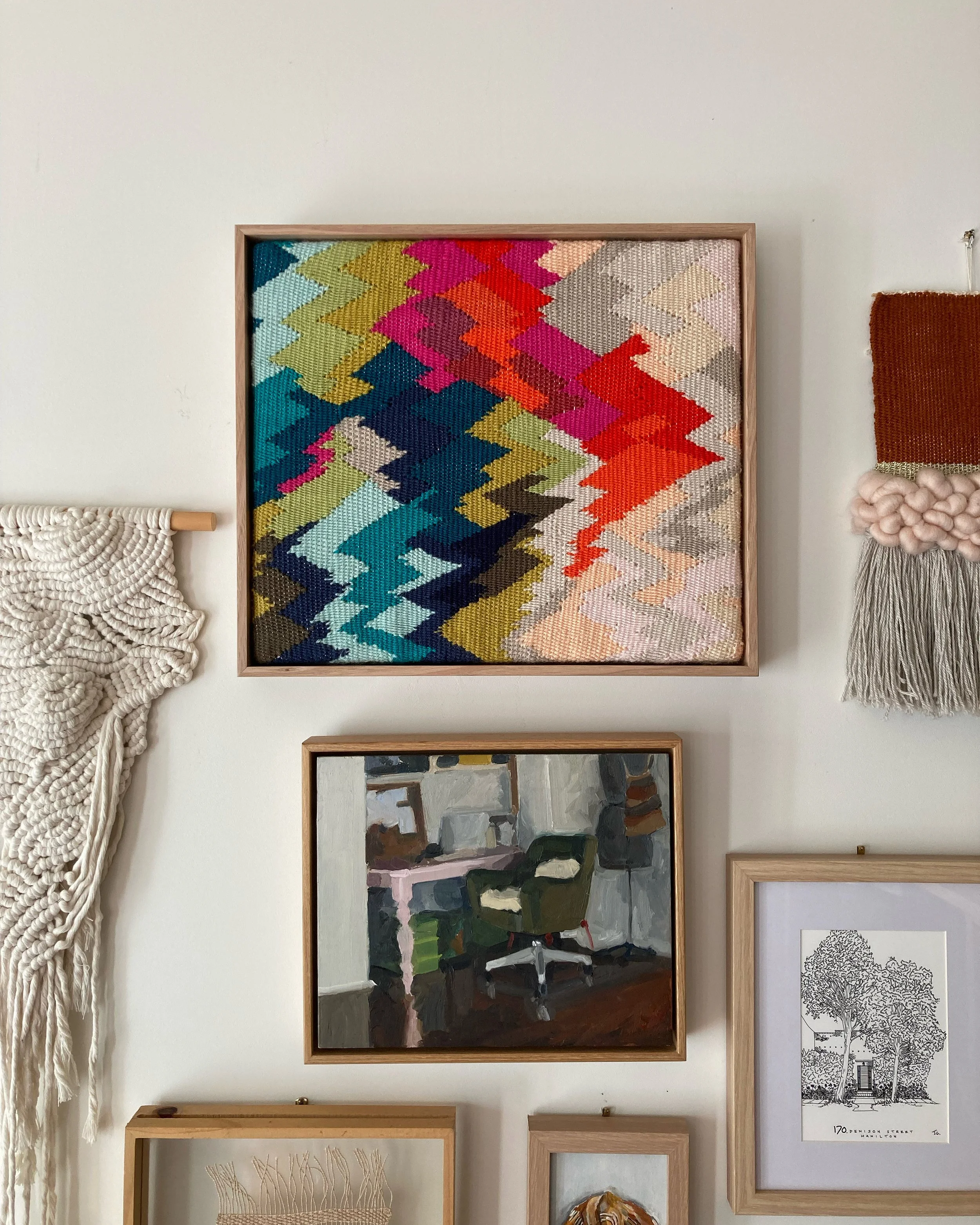 A wall with various framed artworks including a colorful abstract woven piece, a painting of a room with a chair and desk, a black and white sketch of a house and trees, and macramé decorations.