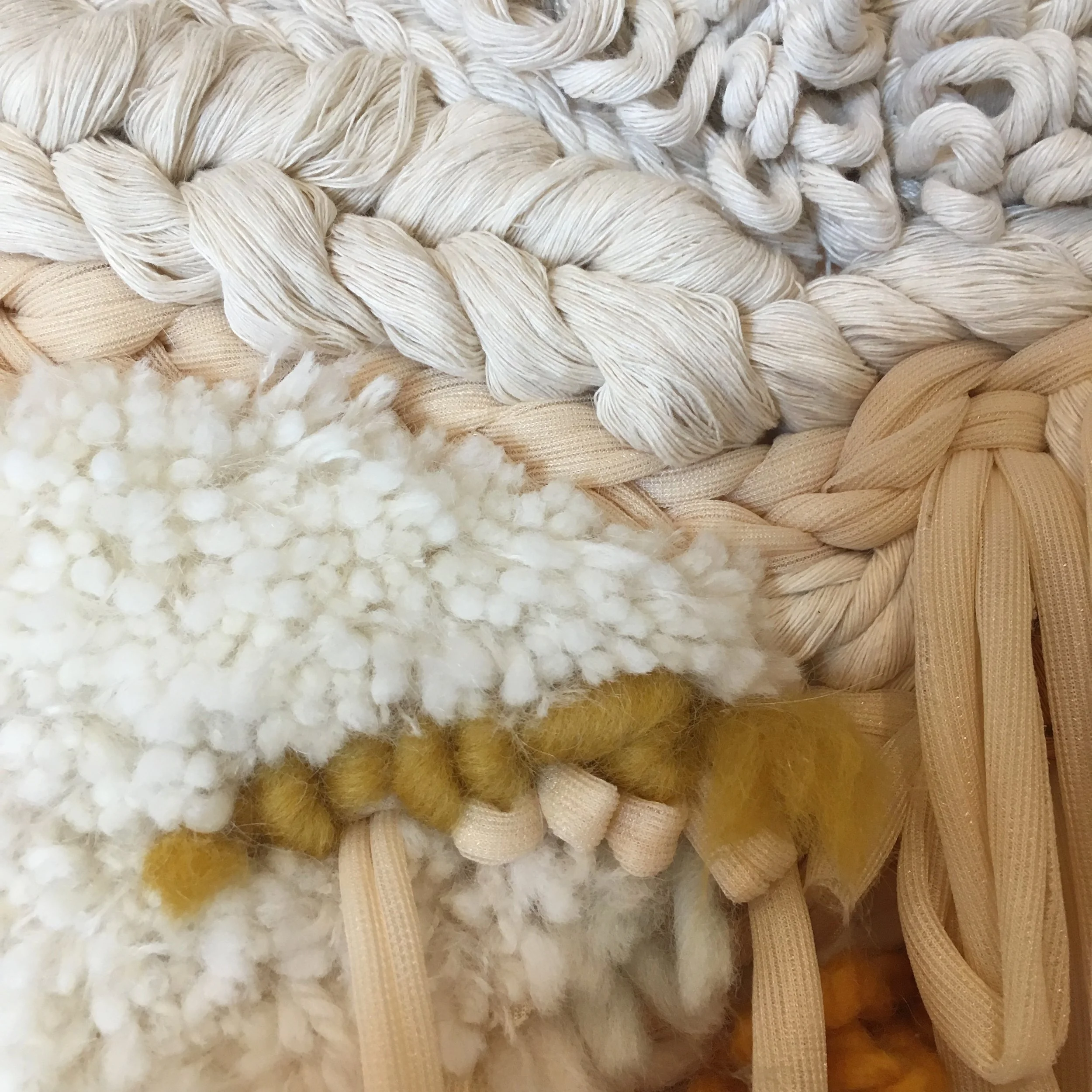 Close-up of a woven textile with different textures, including thick yarn, braided fibers, and soft, fluffy tufts in beige, white, and mustard yellow hues.