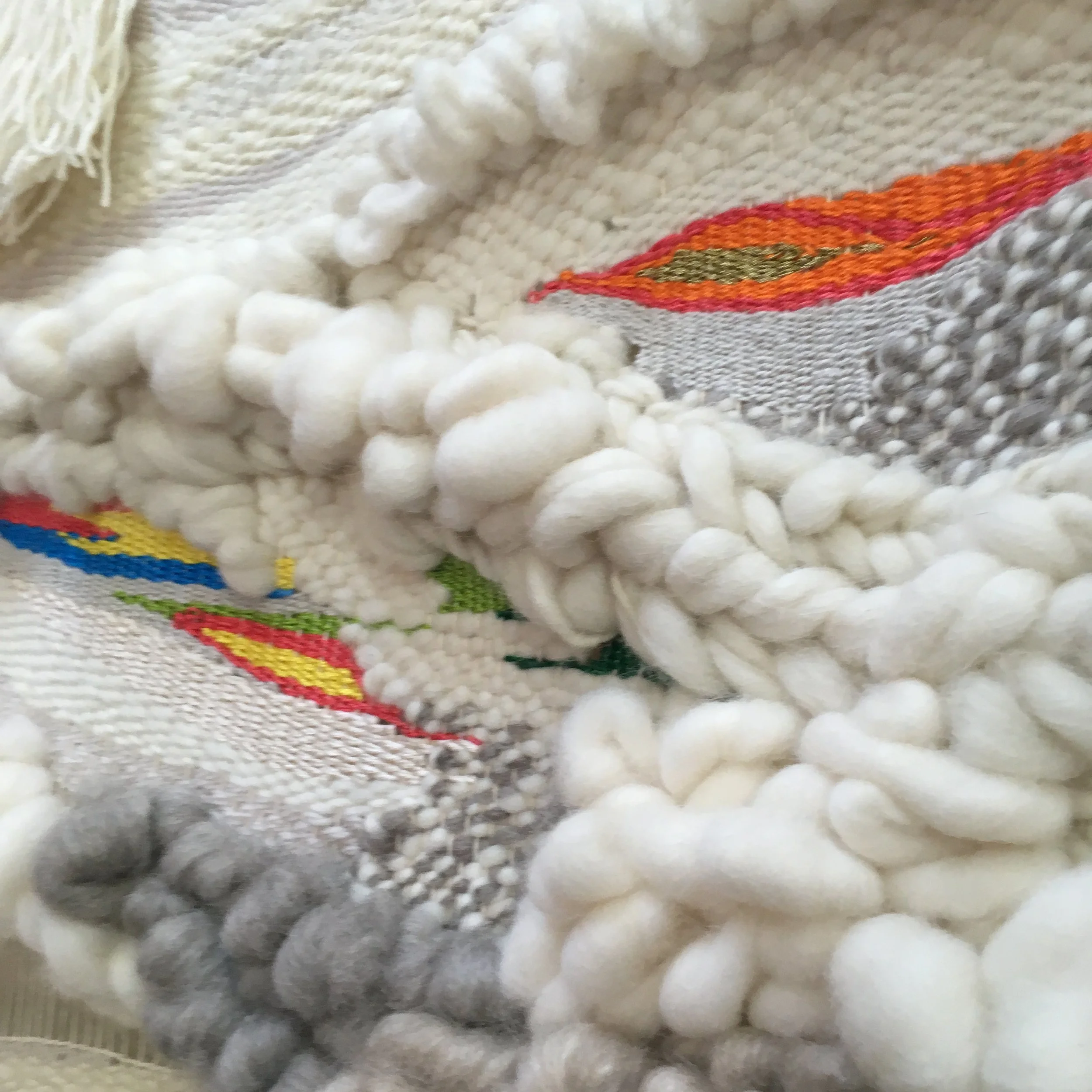Close-up of a textured woven fabric with thick, soft white yarn and colorful thread accents in shades of red, orange, green, and blue.