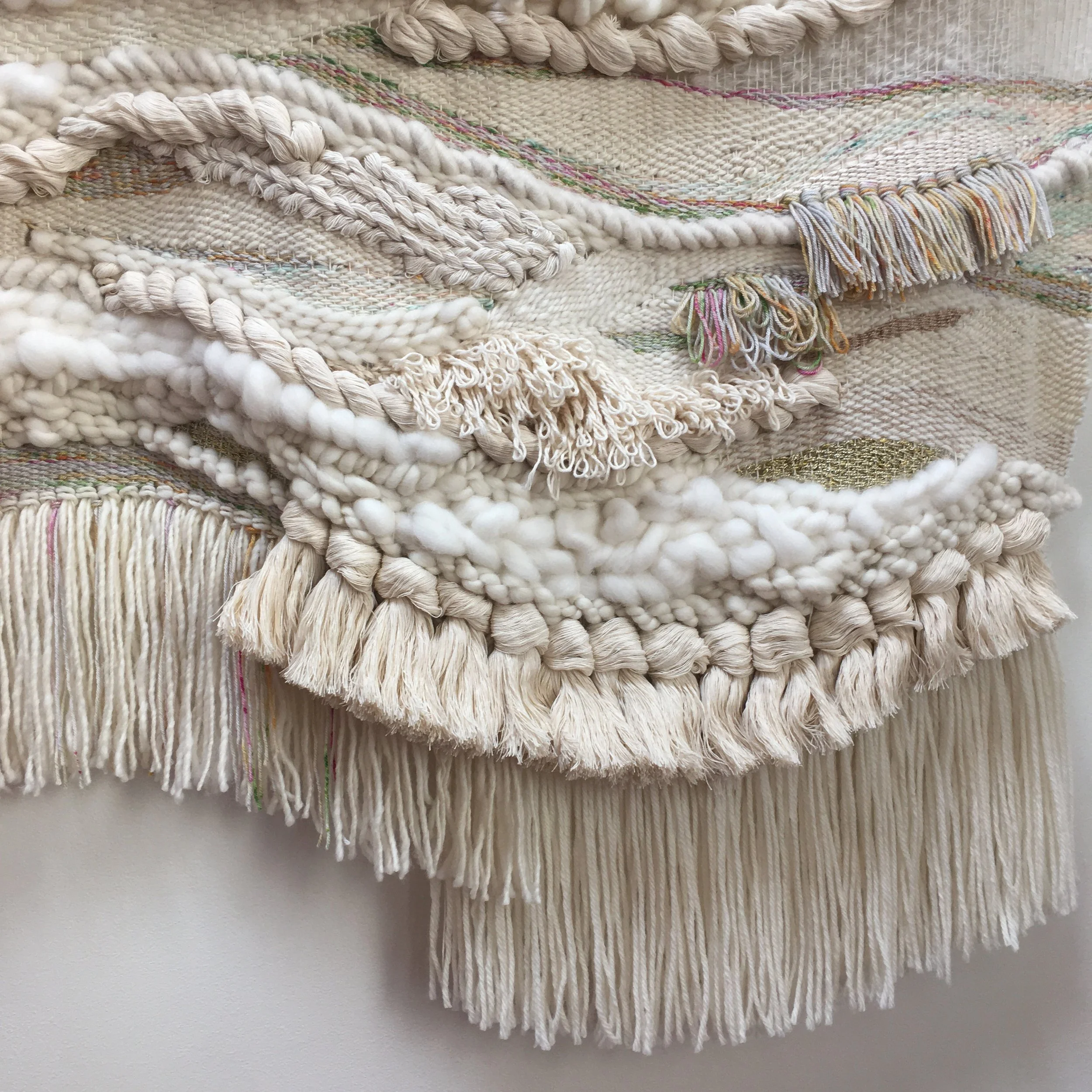 Close-up of a woven textile with textured fibers, featuring layered fringes and various weaving techniques in neutral tones.