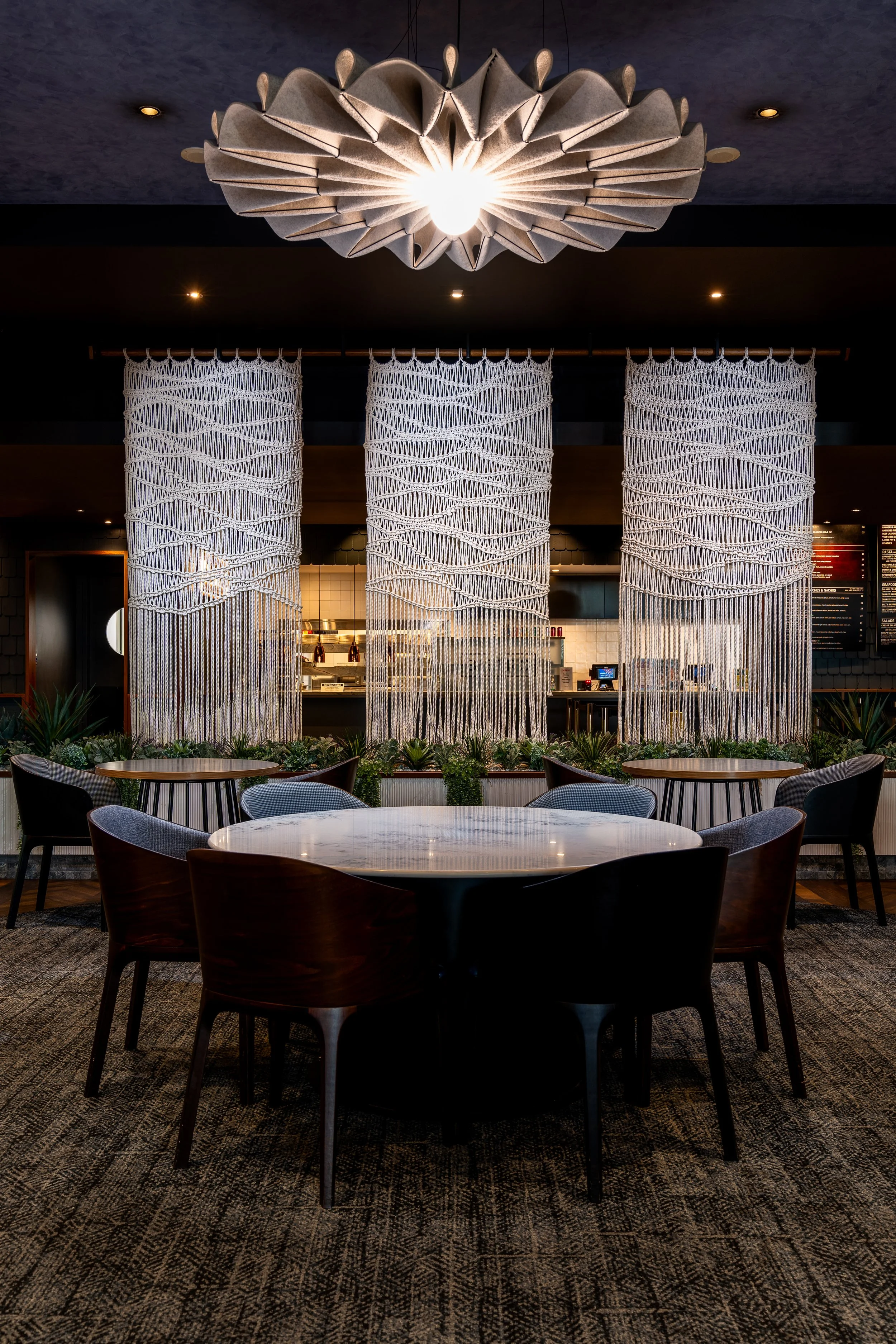 Modern restaurant interior with round tables, chairs, and a decorative ceiling light fixture. Macramé curtains hang above a planter with greenery, and the kitchen area is visible in the background.
