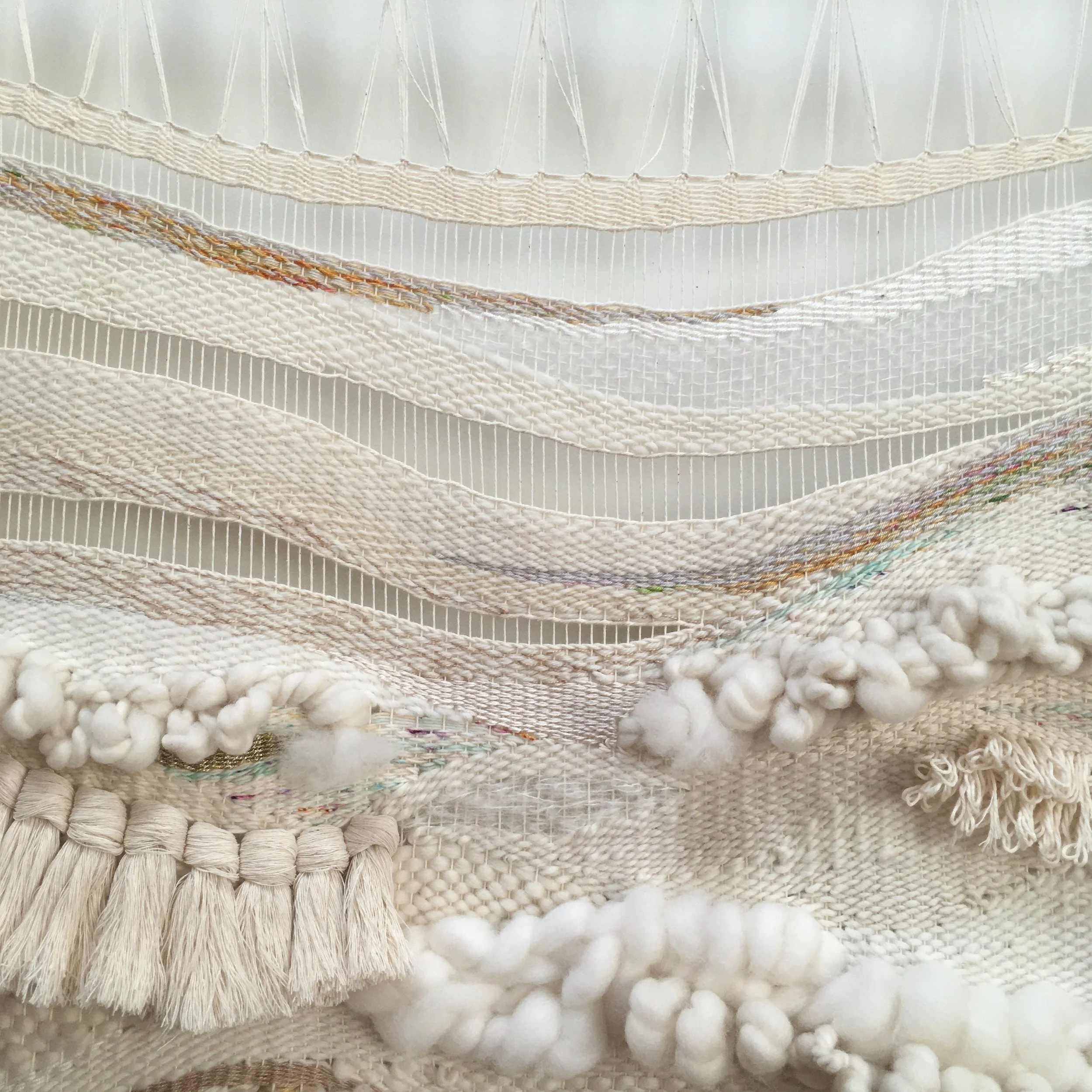 Close-up of a woven textile with various textures and neutral tones, including tassels and knotted yarn.