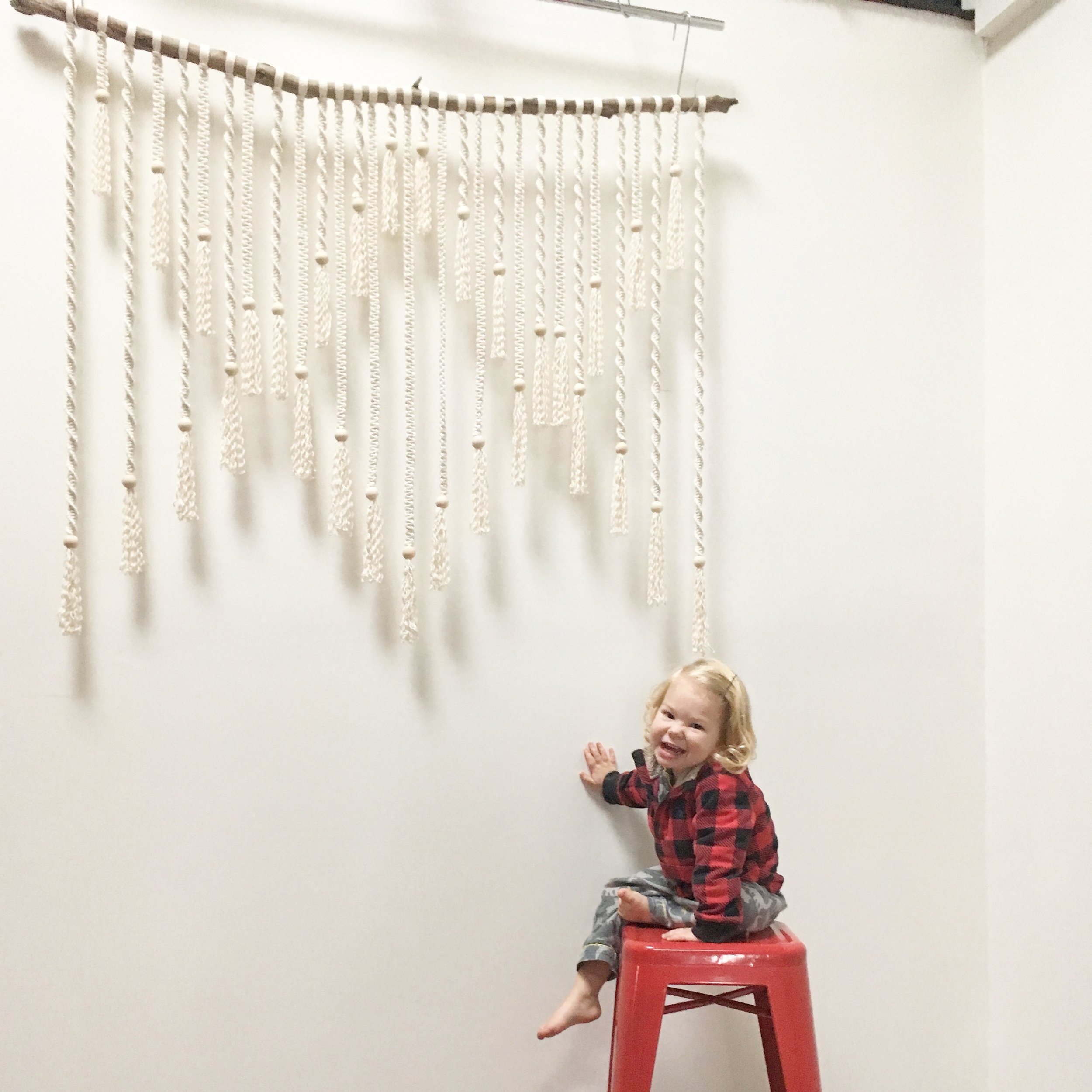 Child sitting on red stool smiling in front of white macramé wall hanging.