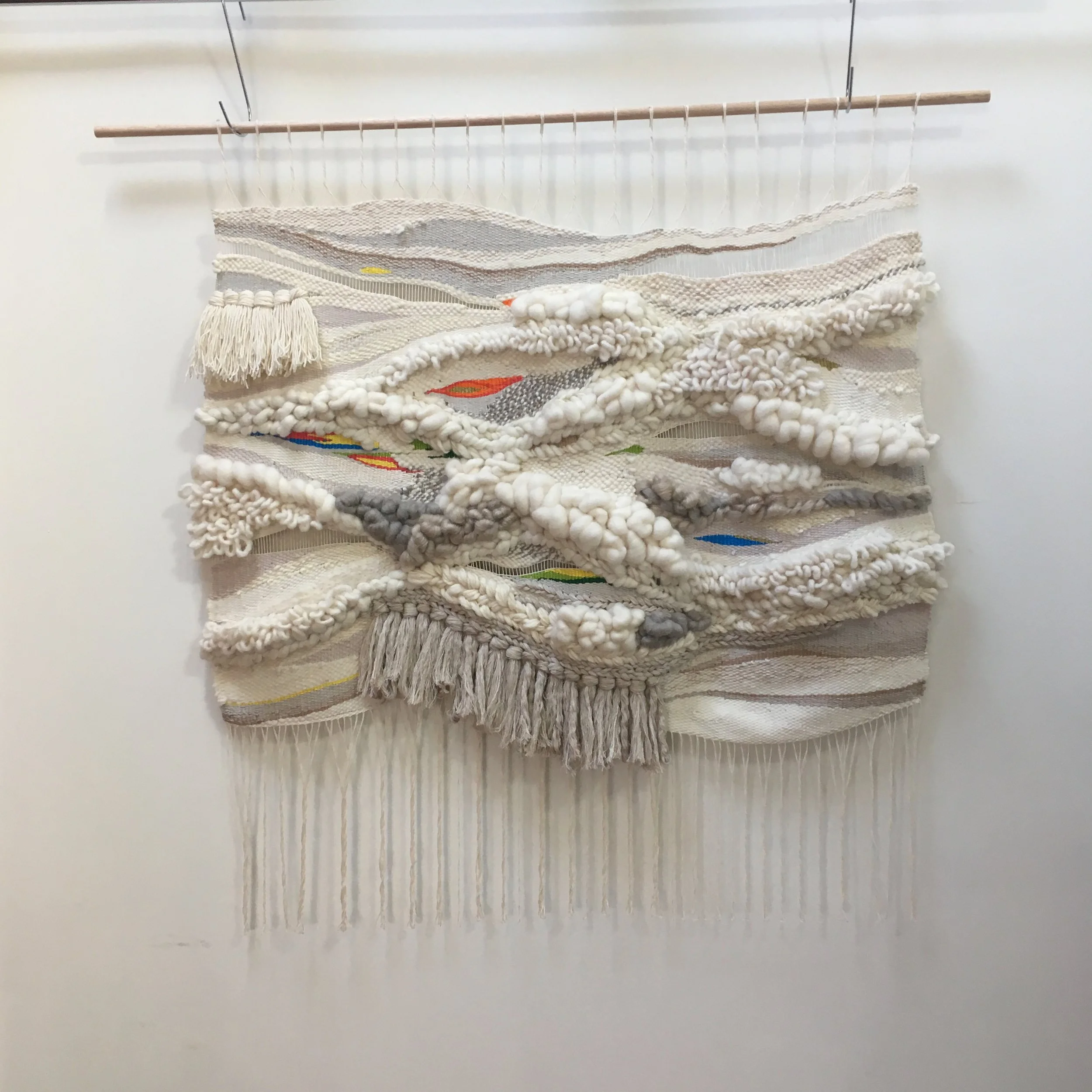Hand-woven textile wall hanging with white and beige yarn, featuring various textures and patterns. Accented with subtle colorful threads and fringes, suspended on a wooden rod.