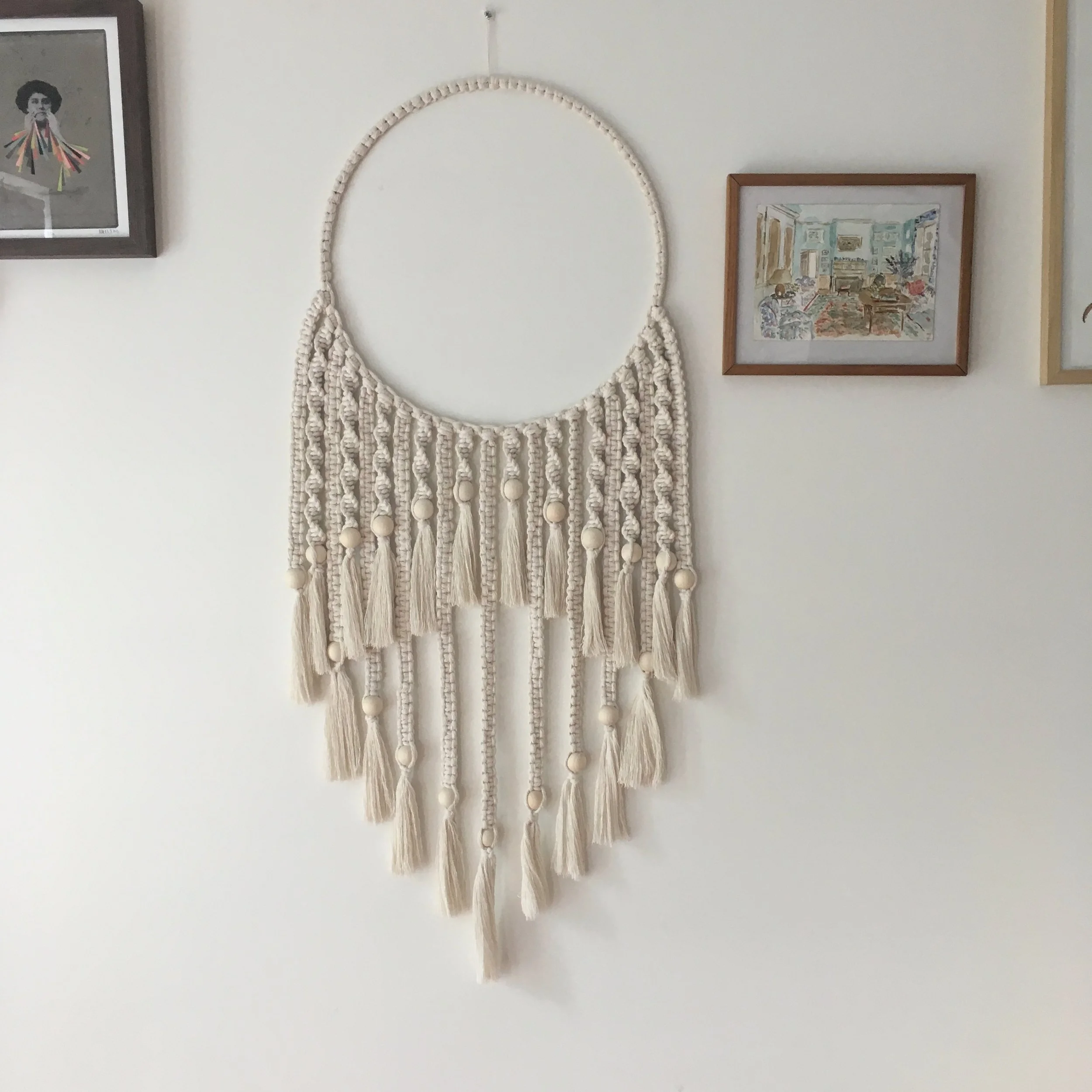 Macrame wall hanging with tassels and beads on a white wall, flanked by two framed pictures.