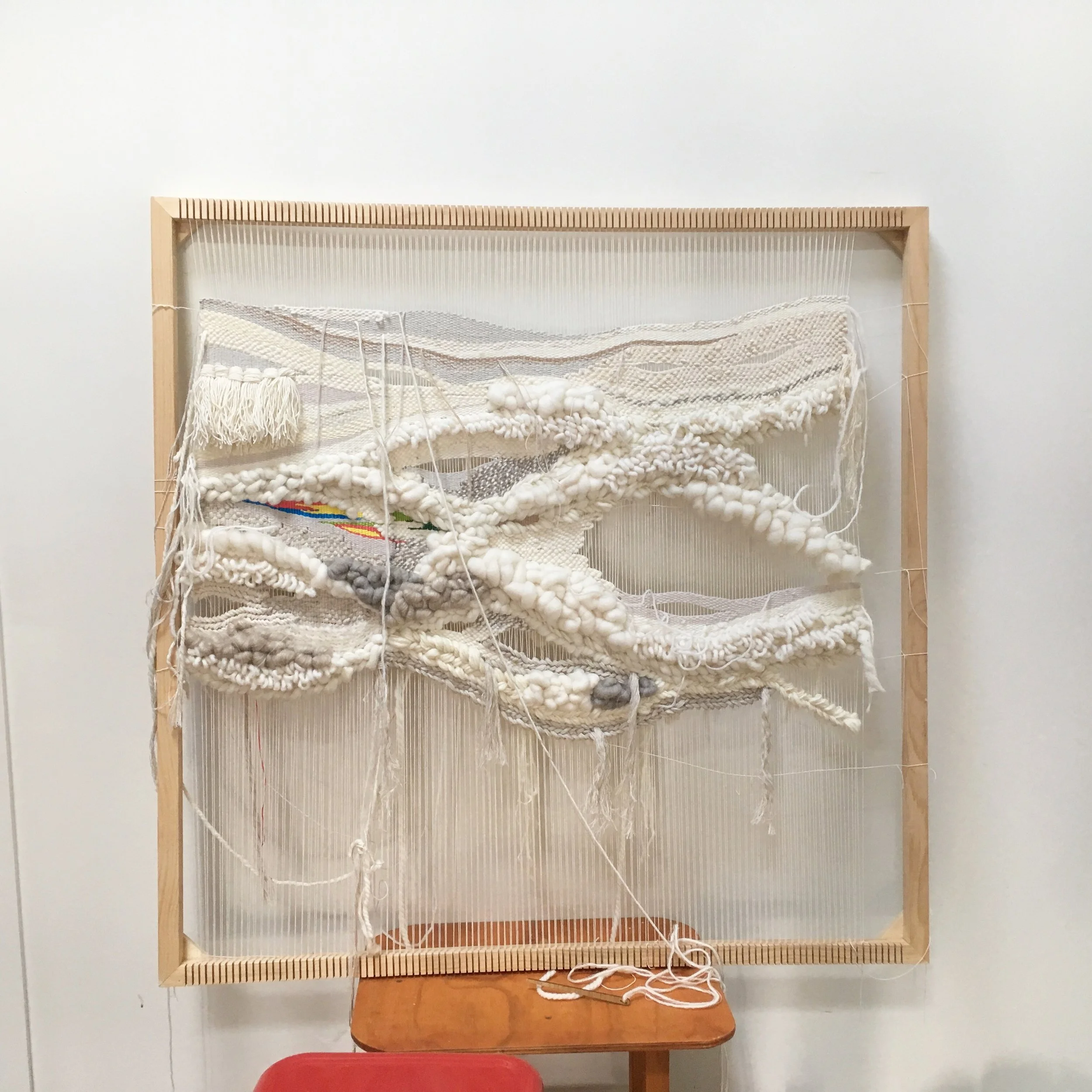Textured wall hanging on wooden frame, featuring white yarn in a woven, layered design, with a small section of colored threads. A chair is partially visible beneath.
