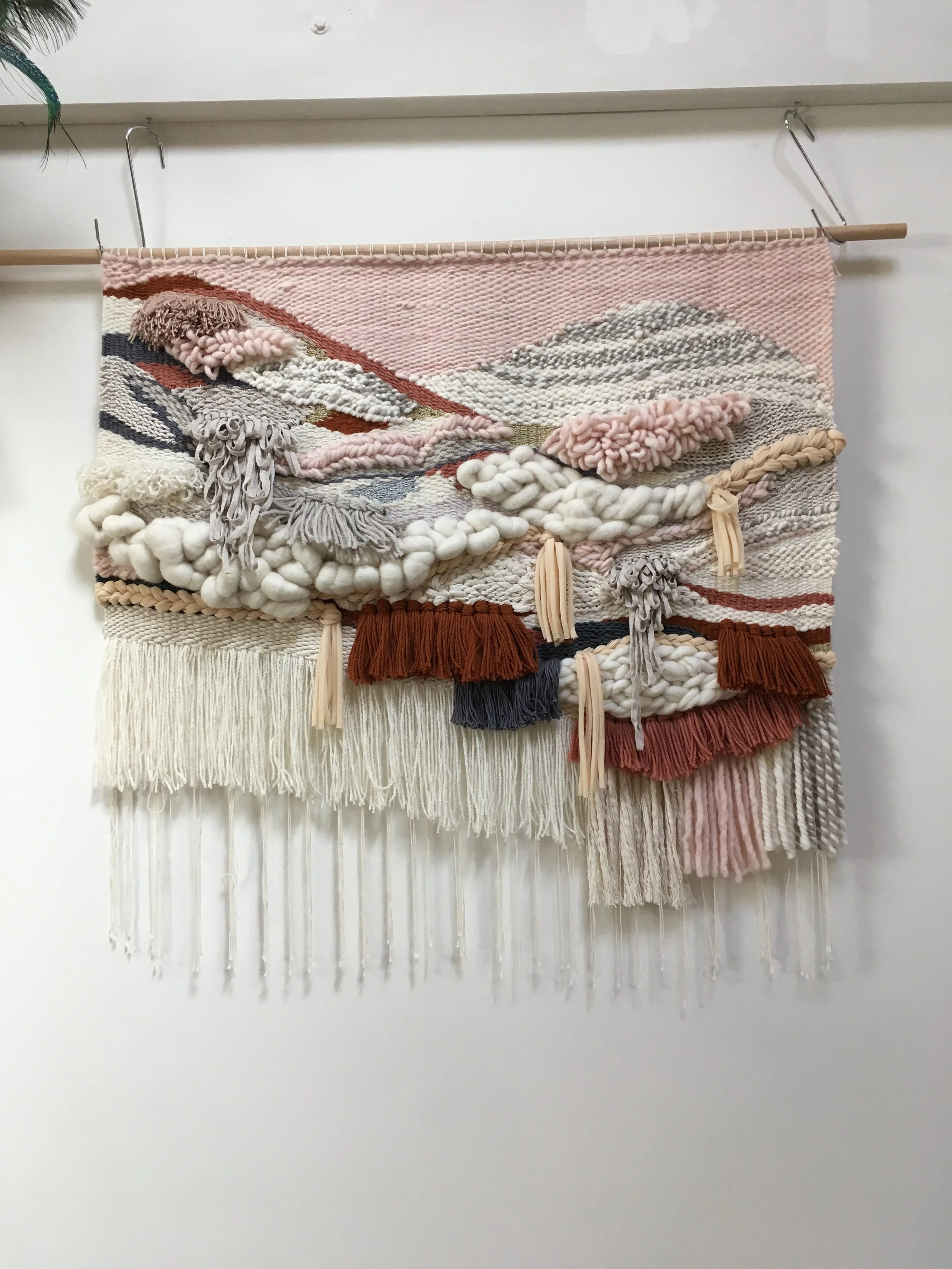 A textured, multicolored woven wall hanging with tassels and fringe, featuring patterns in shades of pink, white, brown, and gray.