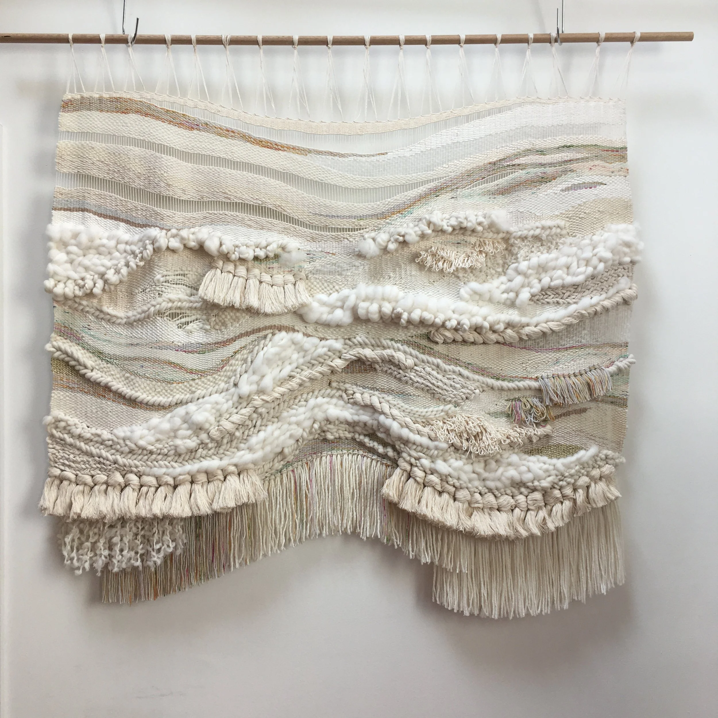 Handwoven textile wall hanging with varying textures and beige tones, featuring fringe detailing and mounted on a wooden rod.
