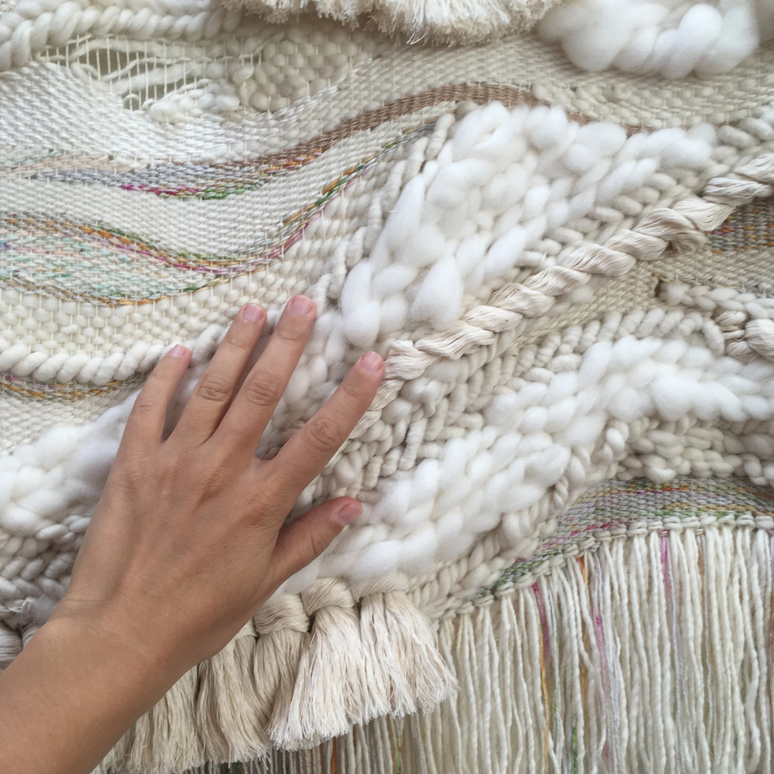 Hand touching textured woven fabric with wool, tassels, and colorful threads.