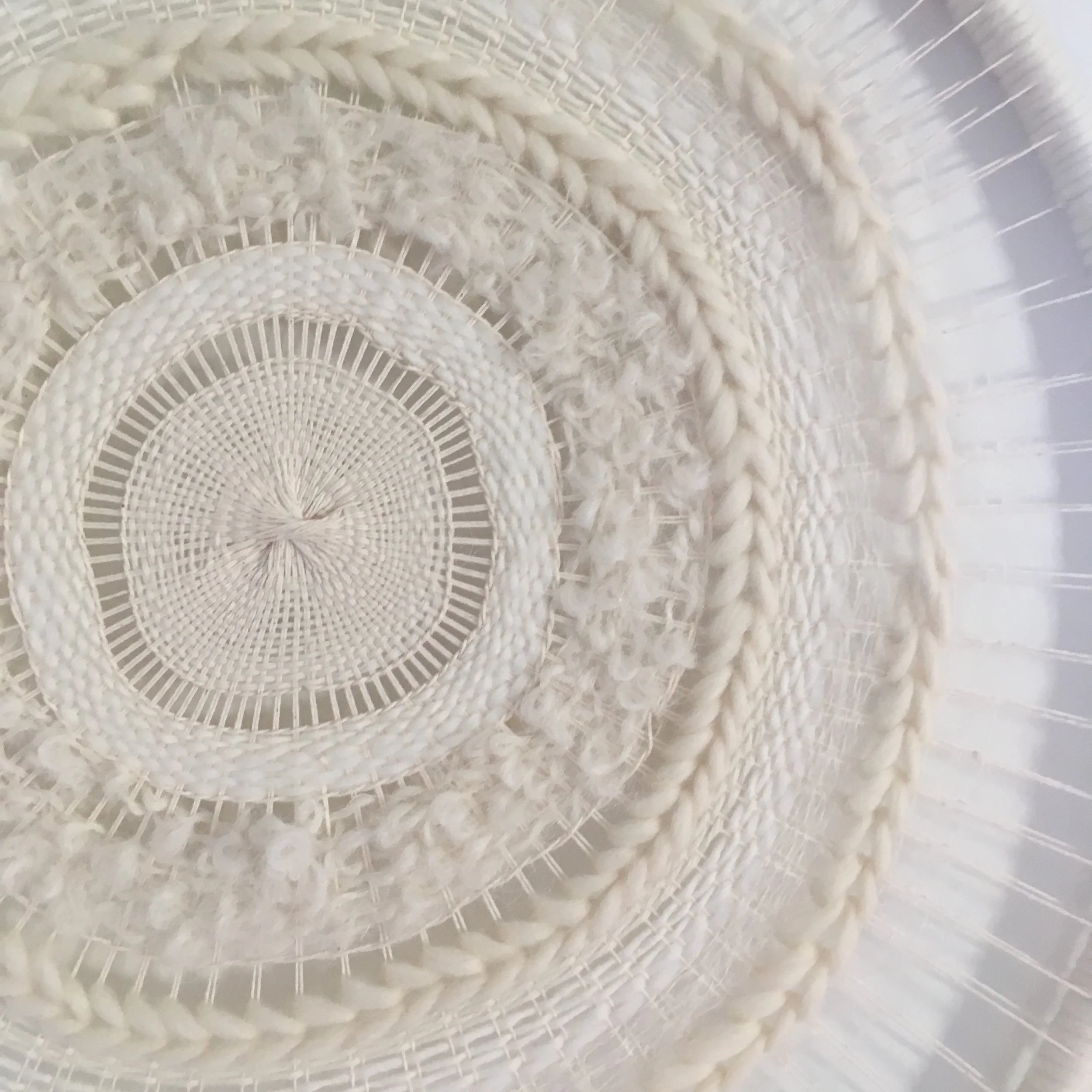 Close-up of a circular woven textile design with concentric patterns in shades of white and cream, featuring various textures and weaving techniques.