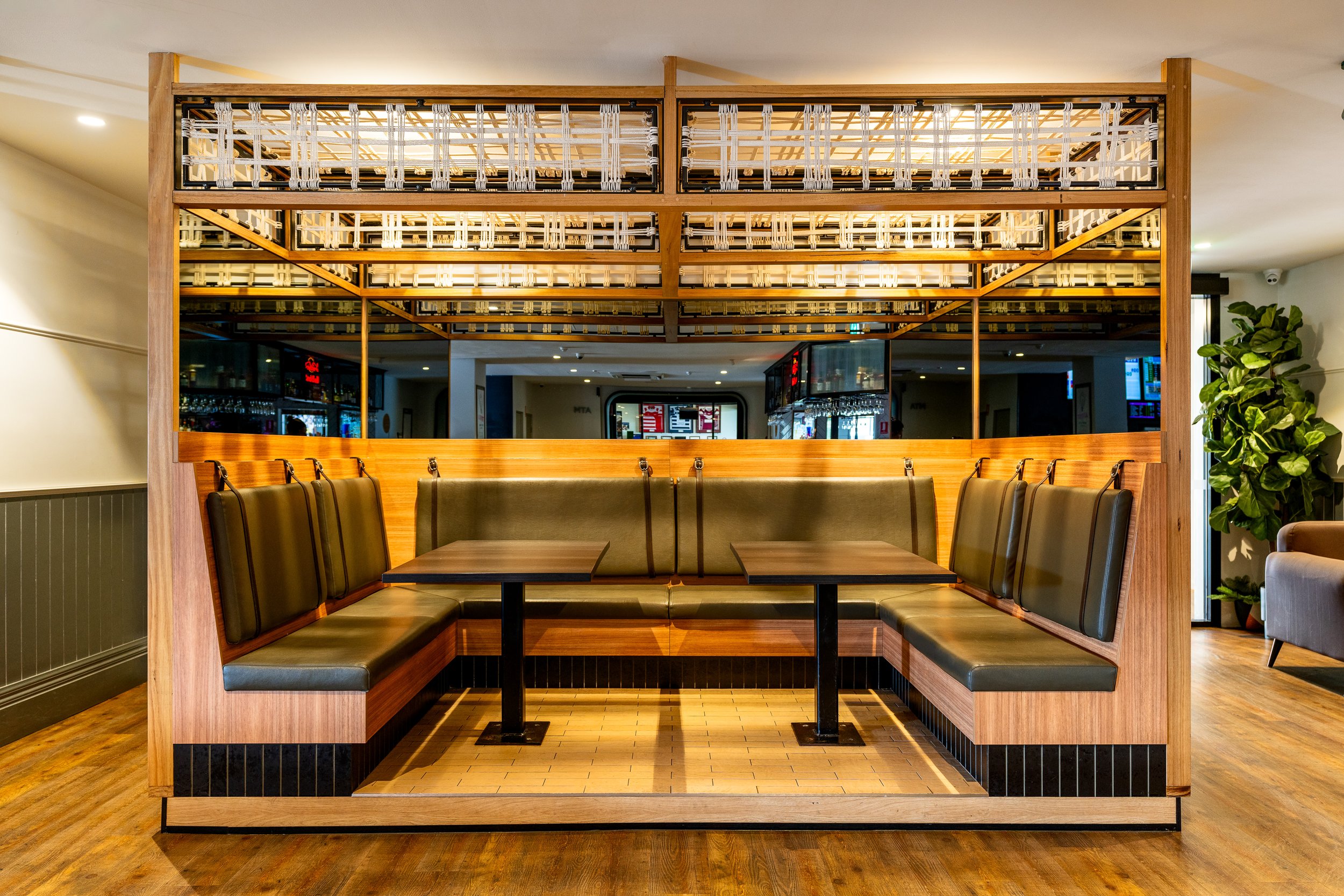 Interior of a modern restaurant with a large booth seating area featuring wooden paneling, cushioned seats, and two tables. The space has a warm lighting ambiance with mirrors and decorative elements overhead. A plant is visible in the background.