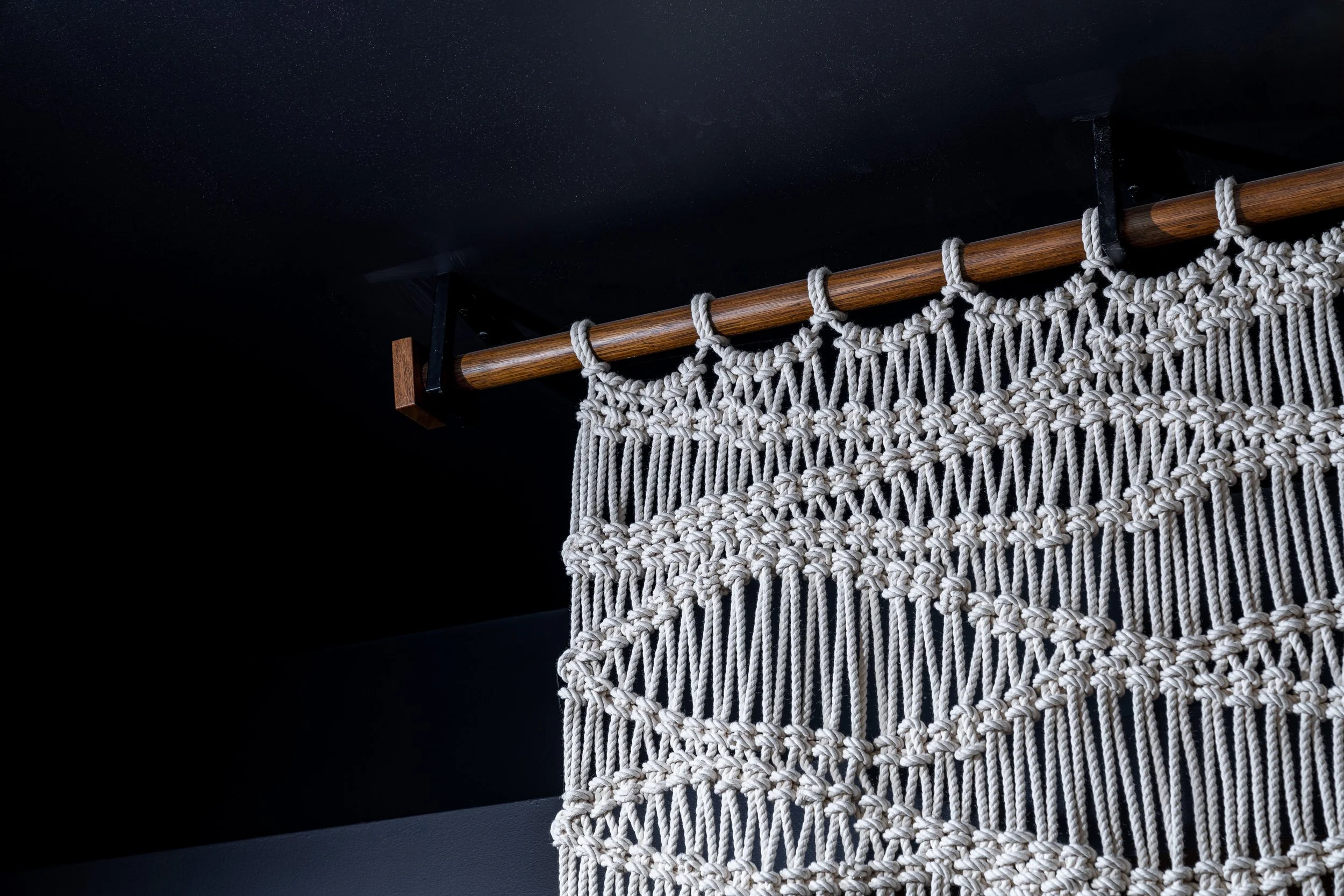 A macramé wall hanging with intricate knot patterns on a wooden rod attached to a ceiling.