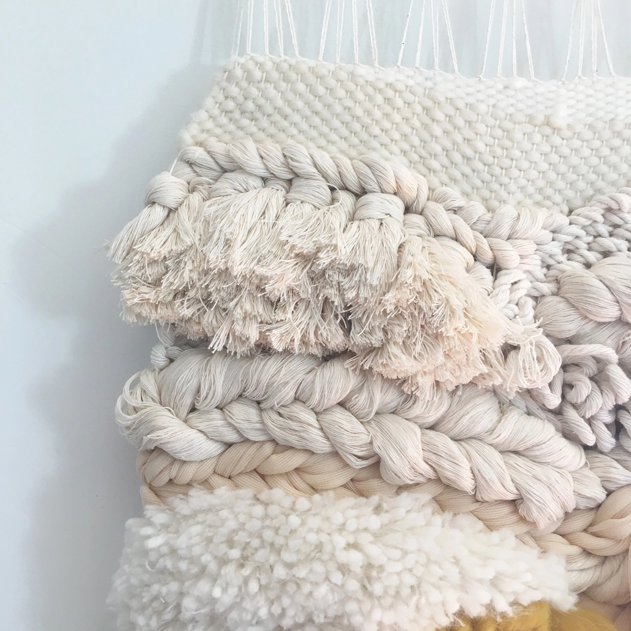 Close-up of textured handwoven wall hanging with various yarns and tassels in neutral colors.