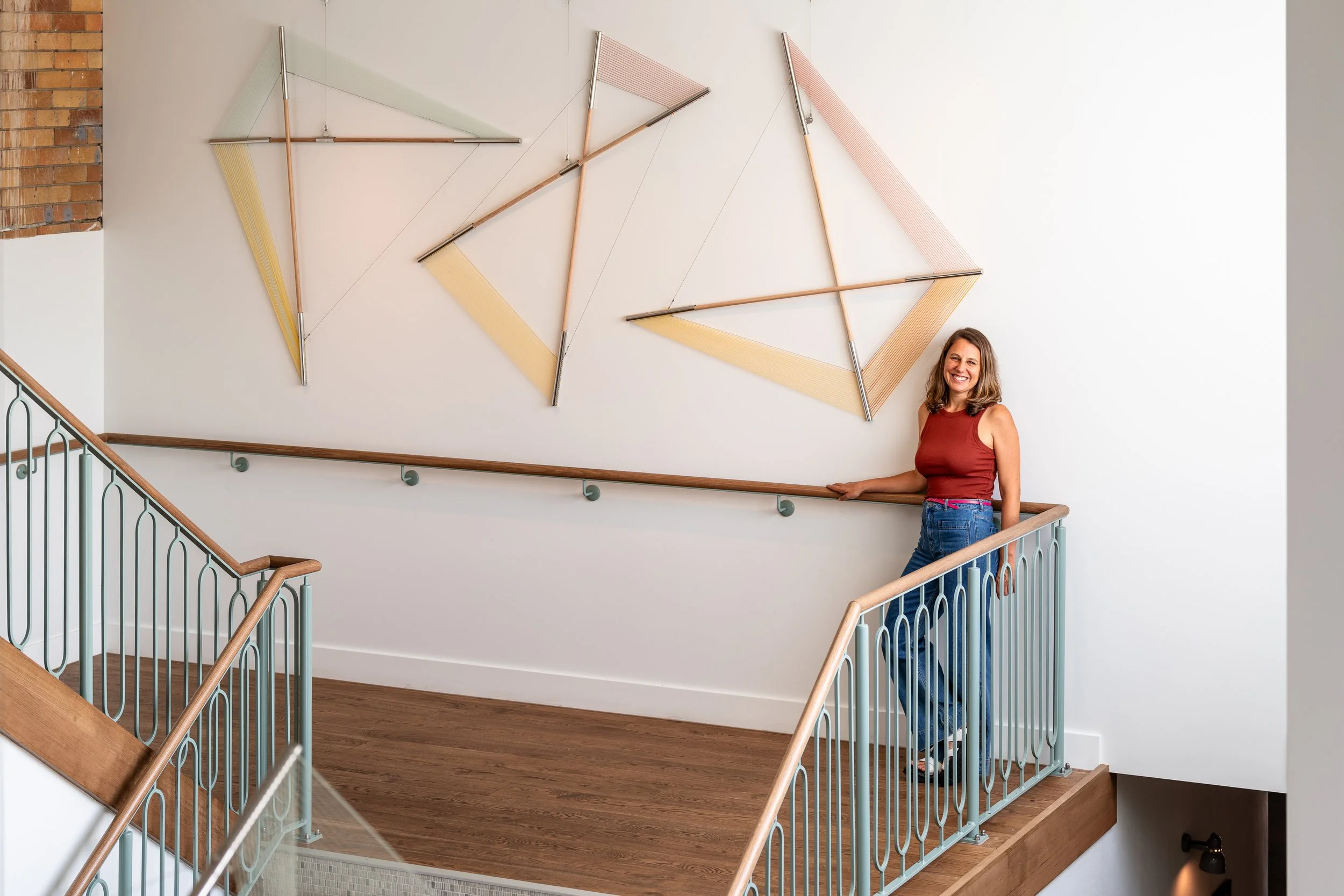 A woman in a red sleeveless top and jeans stands on a wooden staircase. Geometric wall art is displayed on the white wall. The staircase has metal railings with wooden handrails, and a brick wall is partially visible on the left.