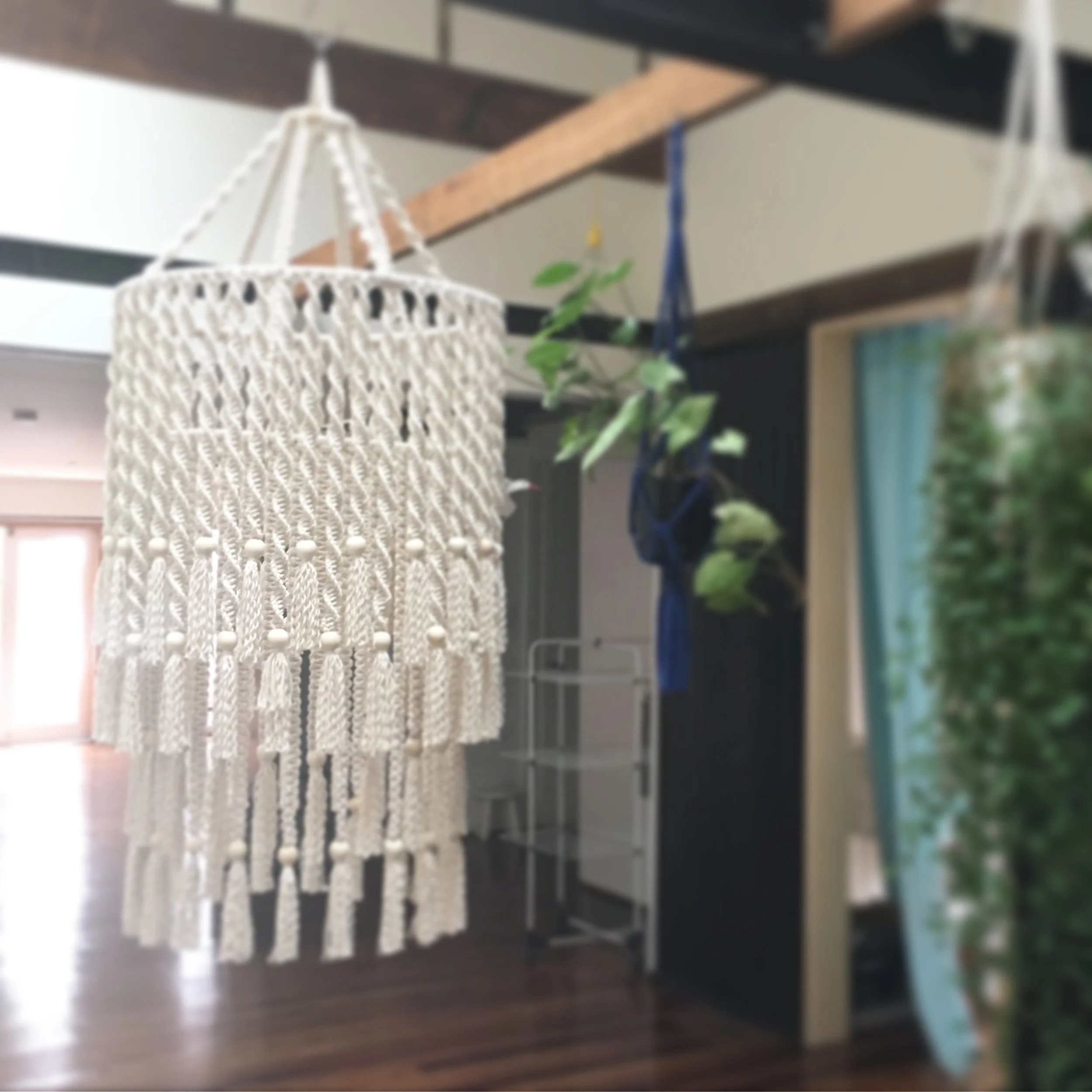Macrame chandelier with tassels hanging from a ceiling in a room with plants and wooden beams.