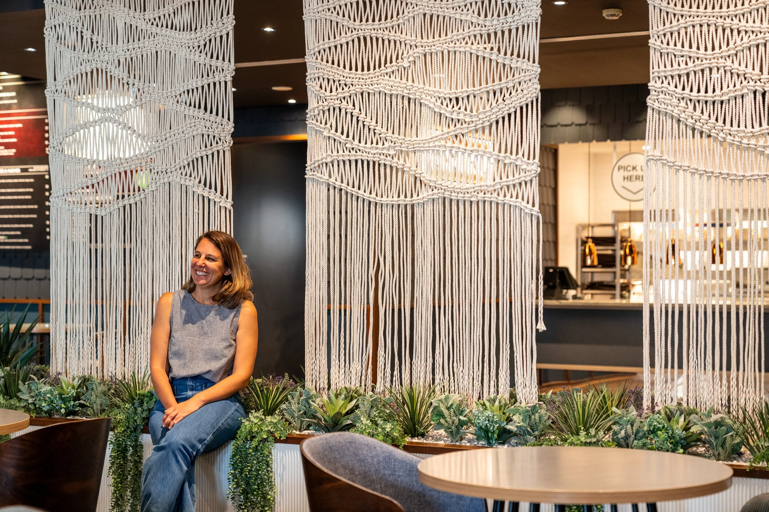 Woman sitting and smiling in a modern cafe with macrame partition and plants.