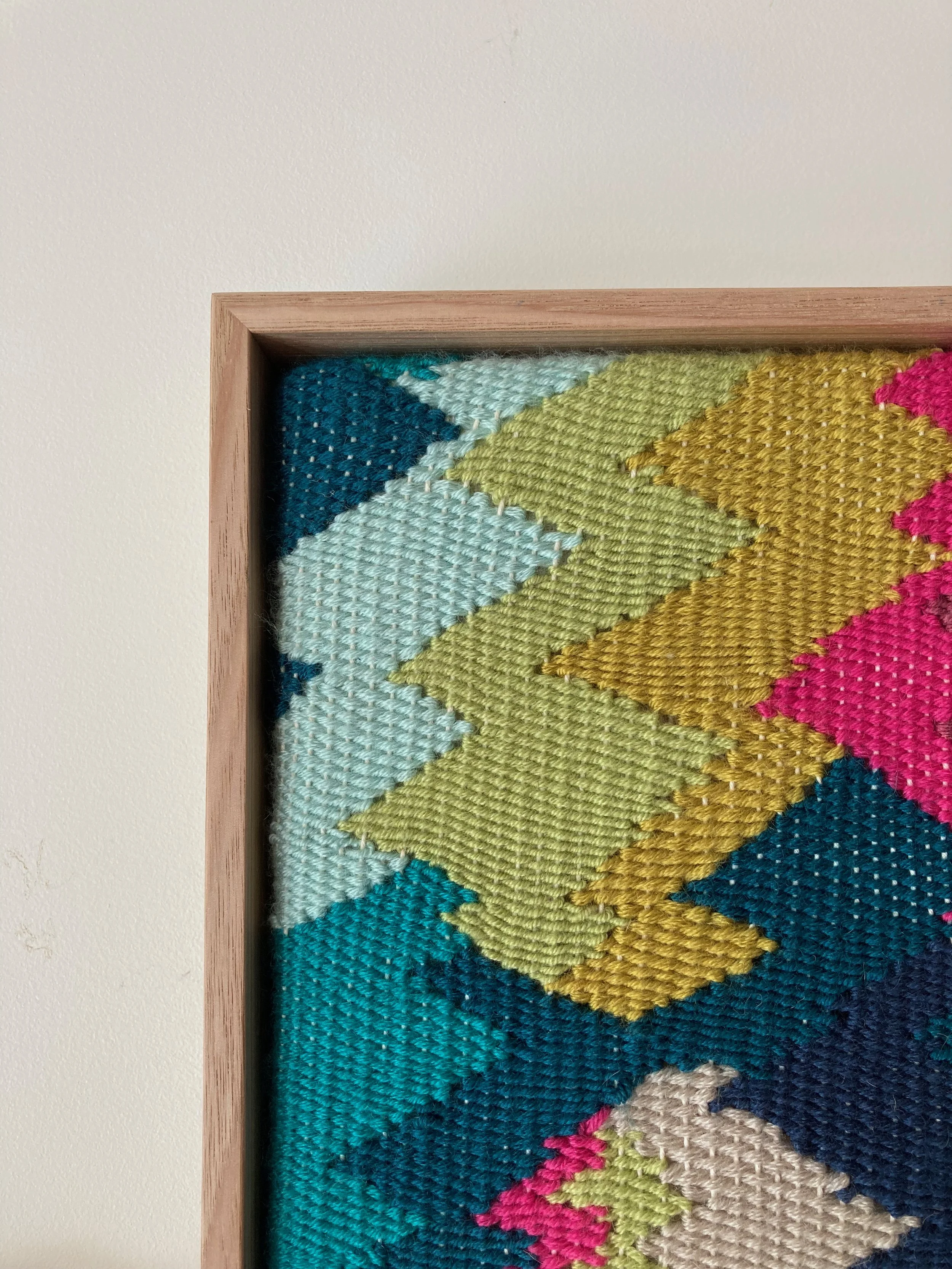 Close-up of a colorful, woven textile framed in wood, featuring a zigzag pattern in blue, green, yellow, and pink hues.