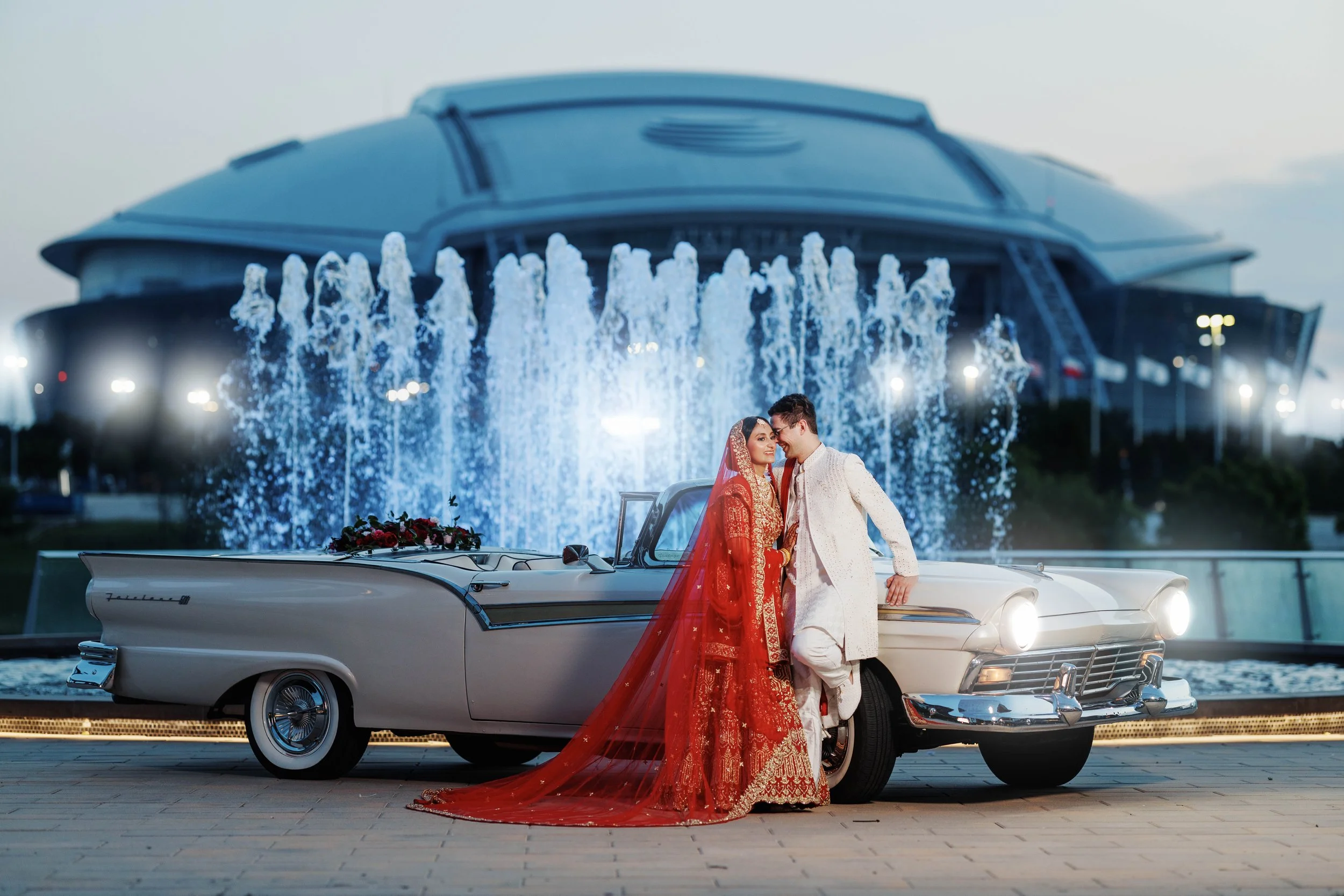 A newlywed couple dressed in traditional Indian wedding attire standing next to a vintage car with a floral decoration, in front of a fountain and a large modern building in the background, during twilight.