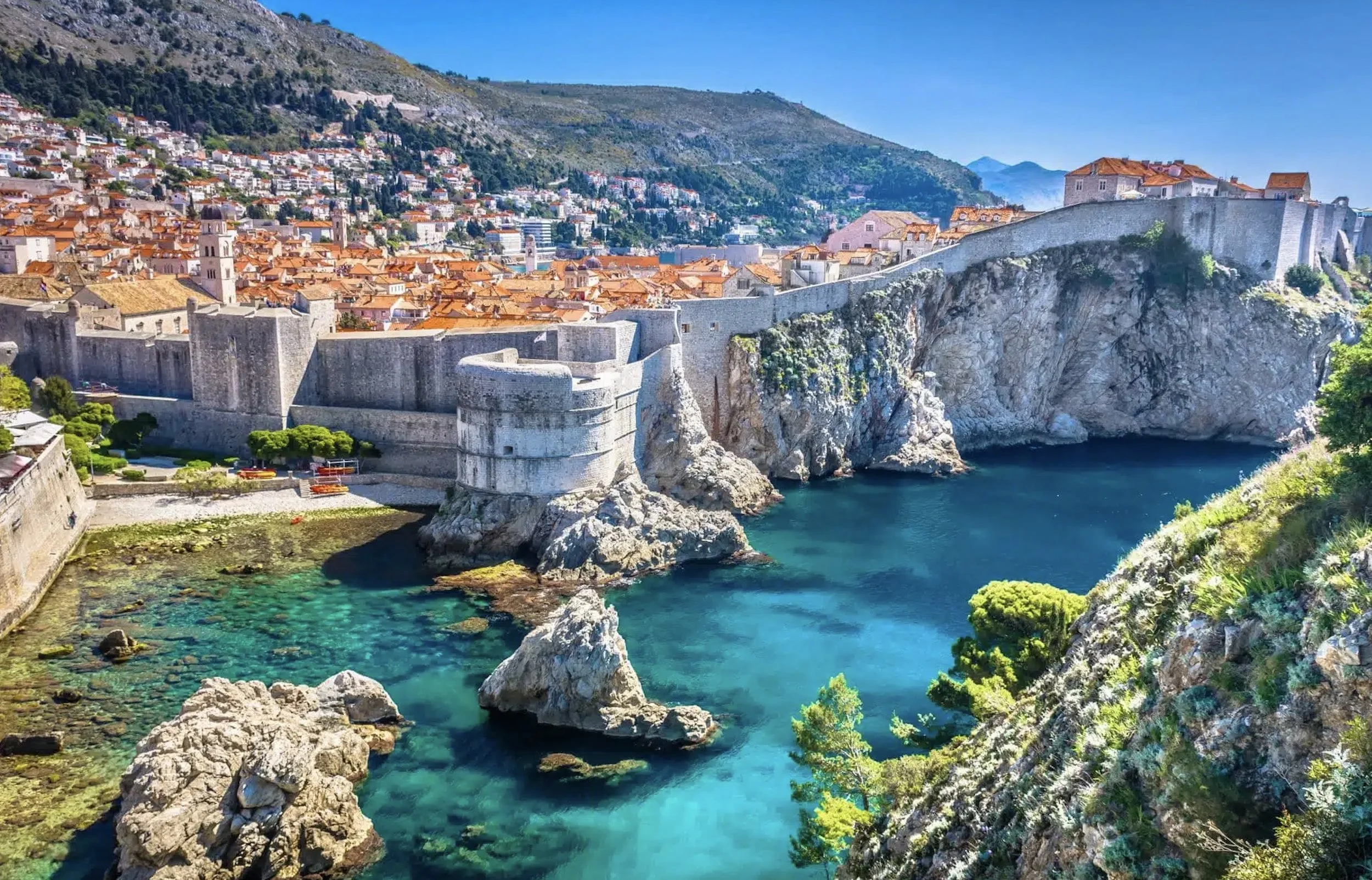 Dubrovnik city view from the sea, with the city walls and orange rooftops visible against a blue sky and mountains.